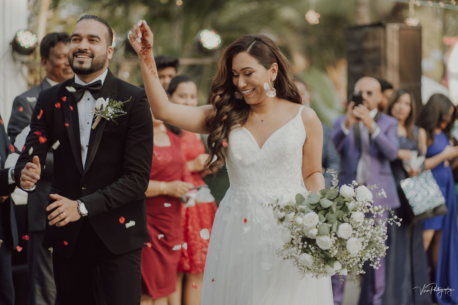 Newlyweds celebrate at outdoor wedding ceremony while guests toss rose petals amid joyful celebration.