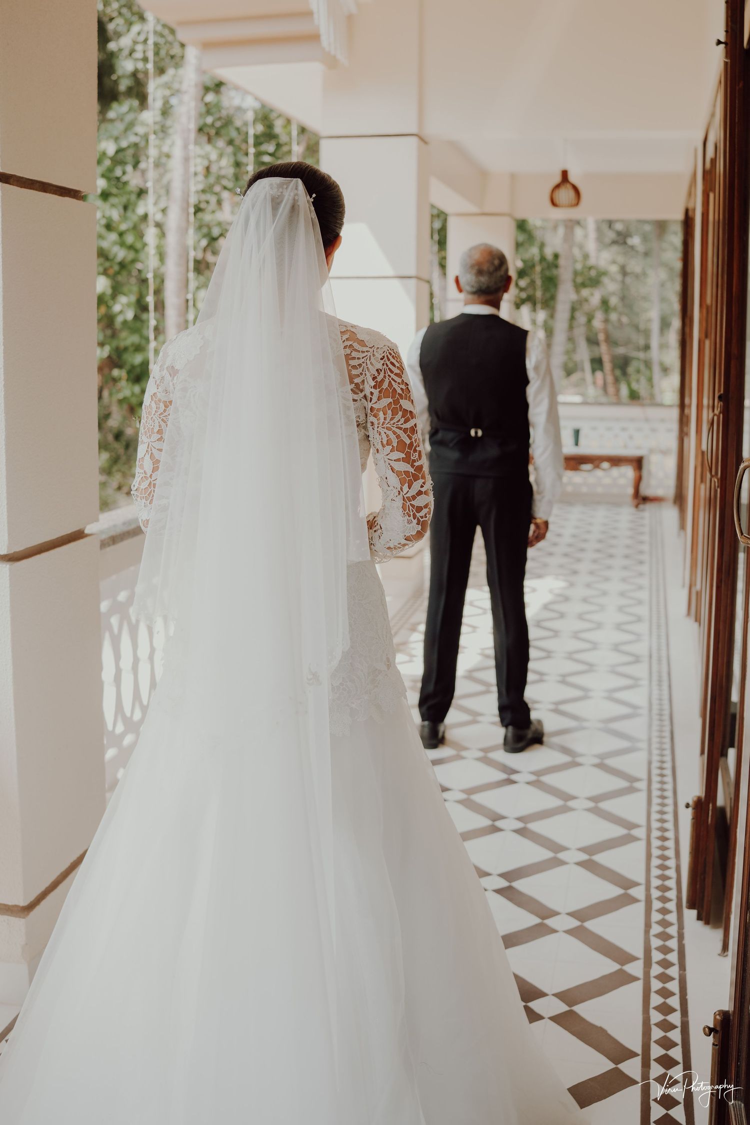 Bride in lace wedding dress with flowing veil stands behind groom in black suit on decorative tiled balcony.