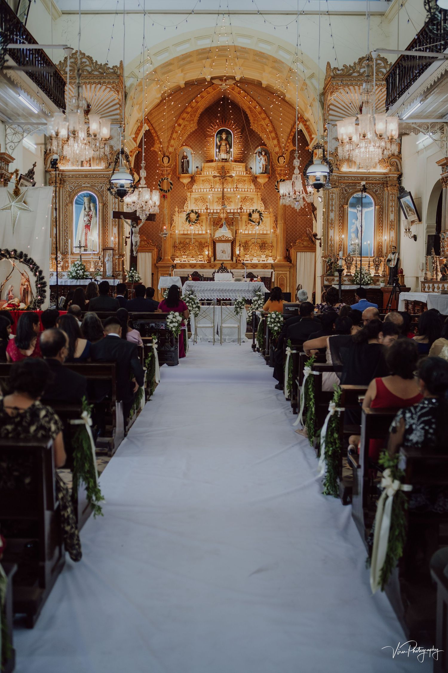 Interior of an ornate Catholic church with chandeliers and white aisle runner during a wedding ceremony.