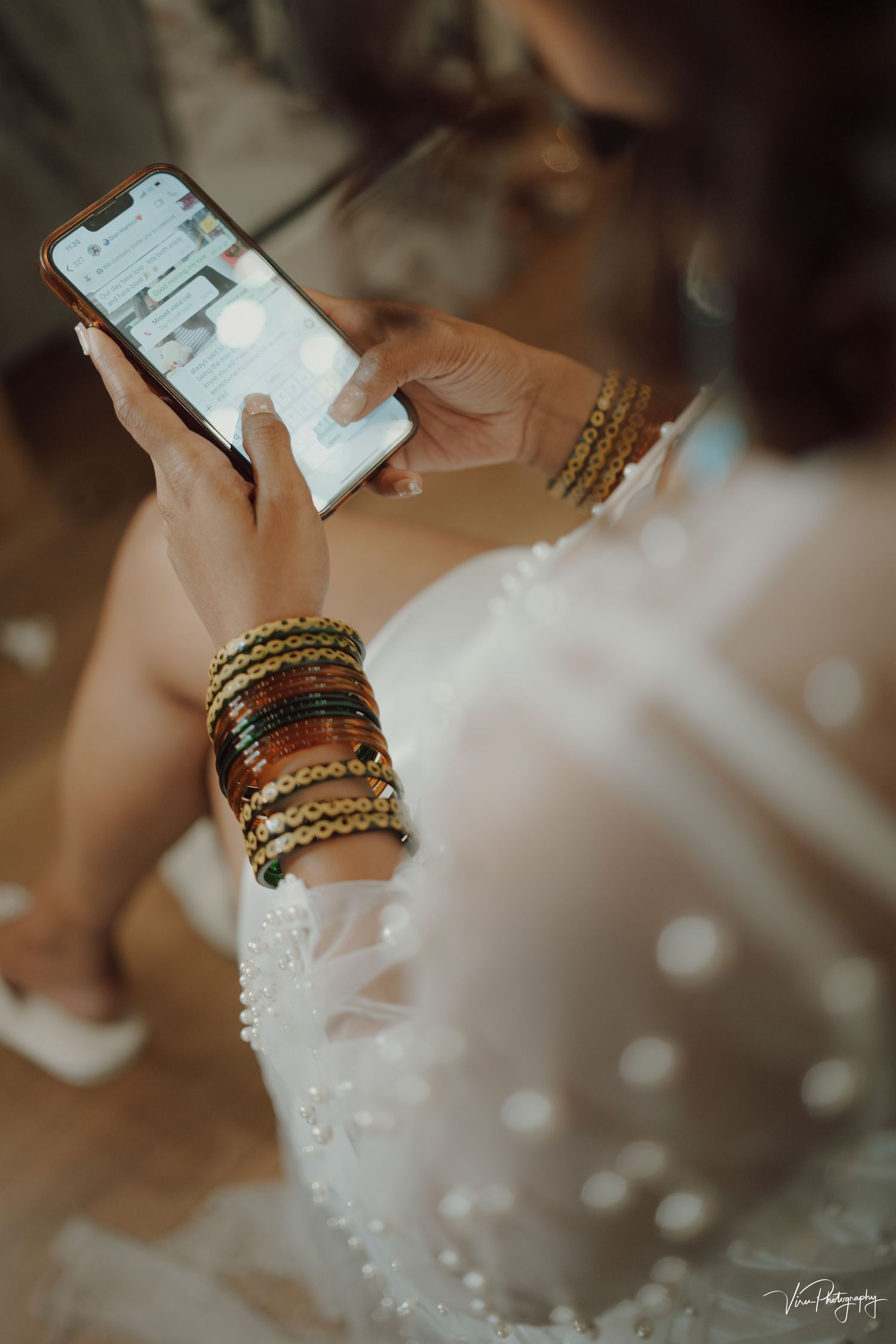 Close-up of hands wearing ornate gold and beaded bangles while checking smartphone in white lace dress.