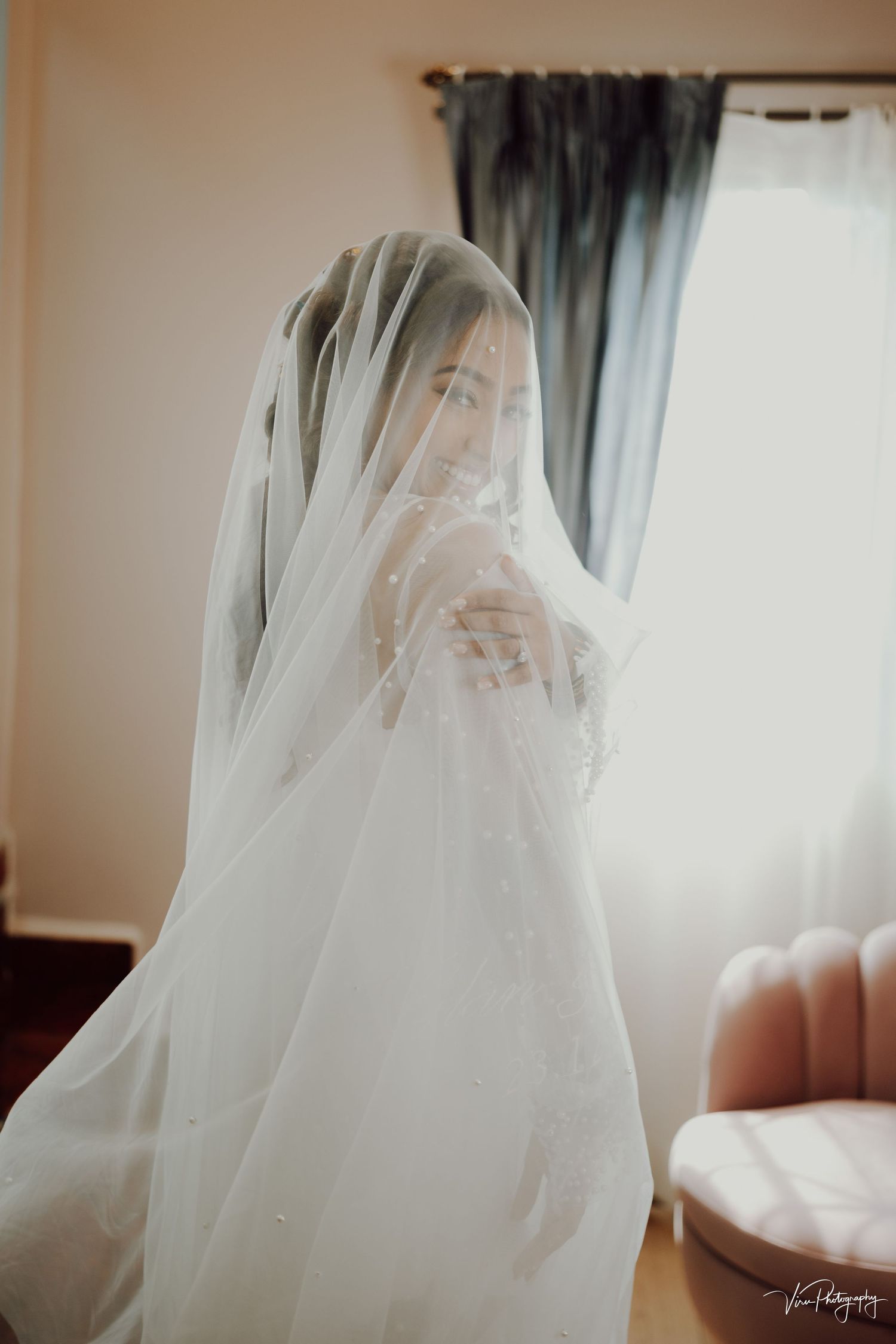 A bride wearing a flowing white veil stands in soft window light creating an ethereal silhouette.