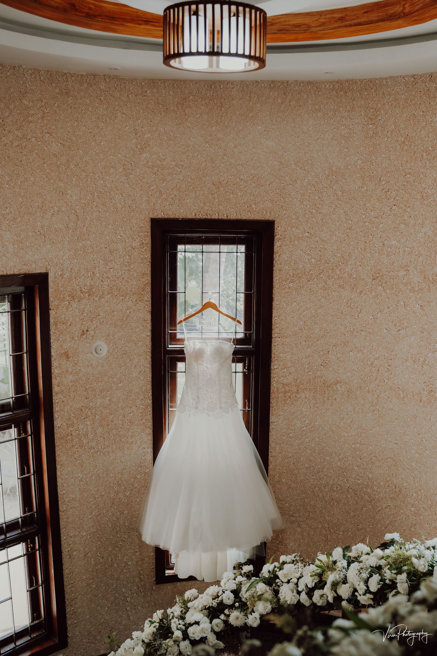 A white wedding dress hanging on a decorative window with a cylindrical pendant light above and white flowers below.