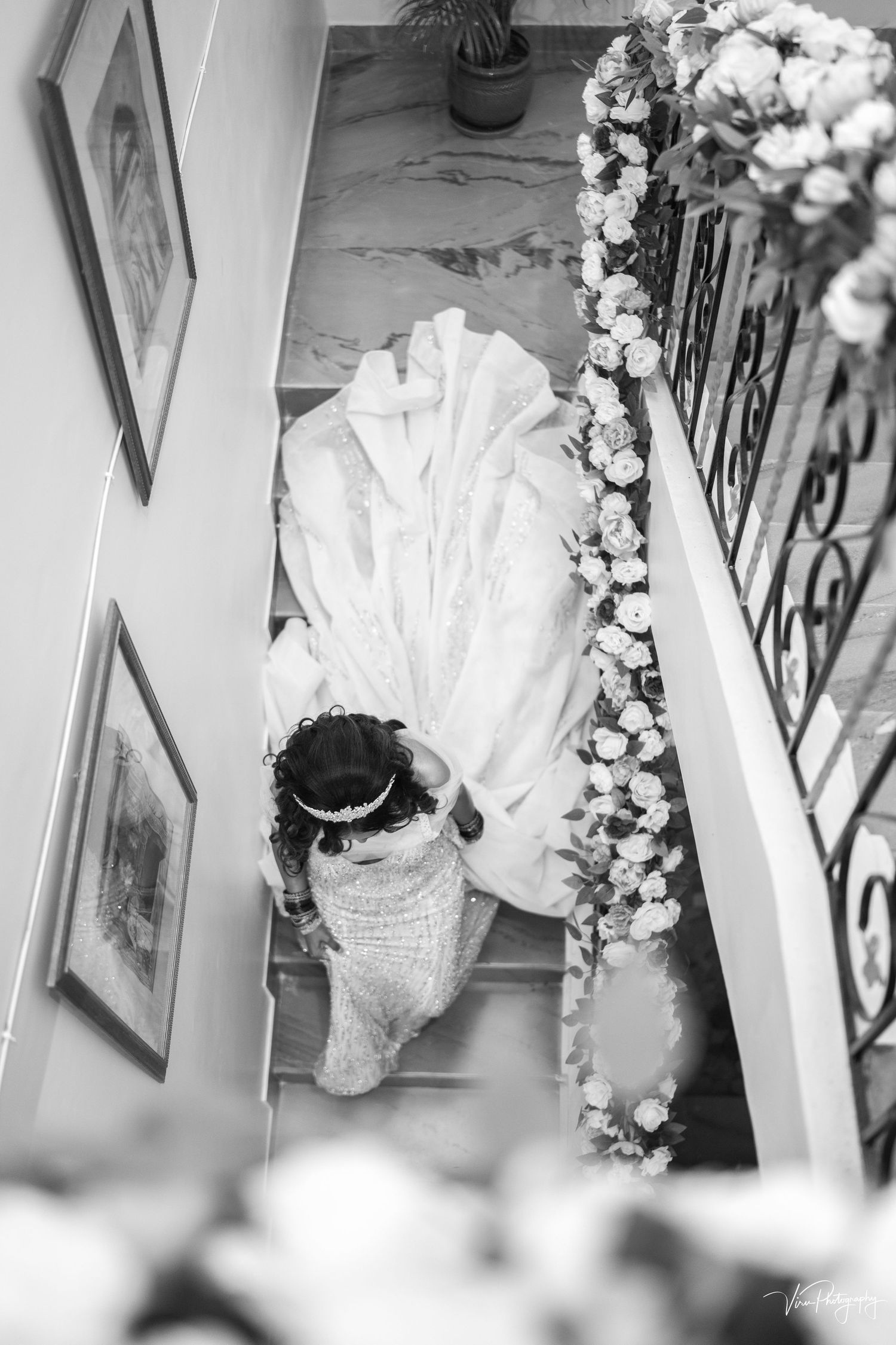 Black and white overhead view of a bride in a flowing white dress descending a floral-decorated staircase.