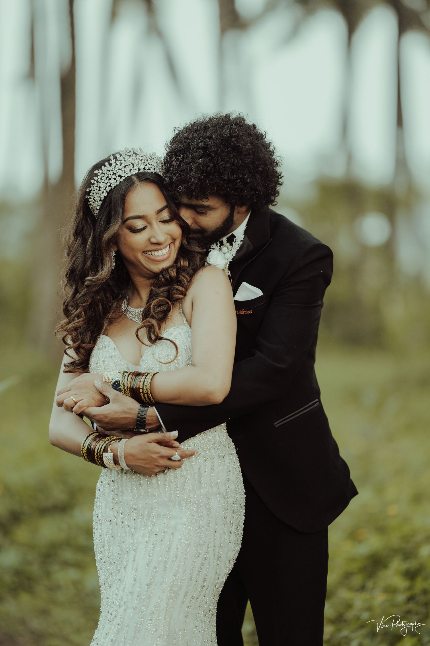 A romantic wedding couple embraces in a dreamy outdoor setting with the bride wearing a beaded gown and crystal headpiece.