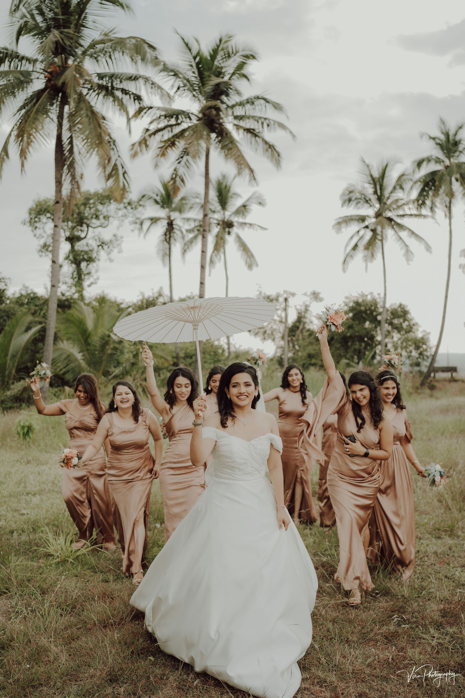 A wedding party with bridesmaids in champagne dresses and a bride in white posing joyfully among tall palm trees outdoors.
