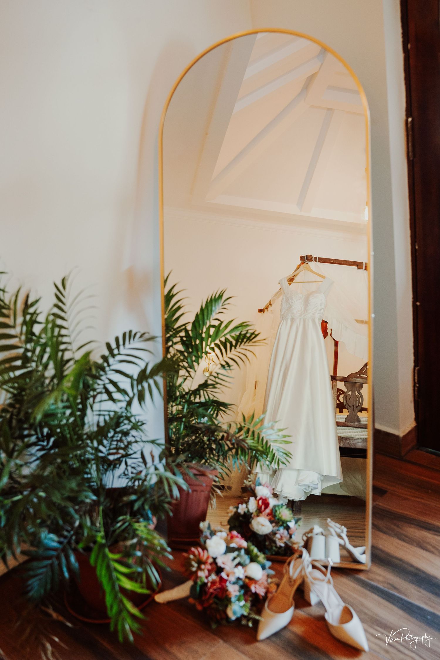 A white wedding dress hangs on a tall gold arched mirror with tropical plants and flowers arranged at its base.