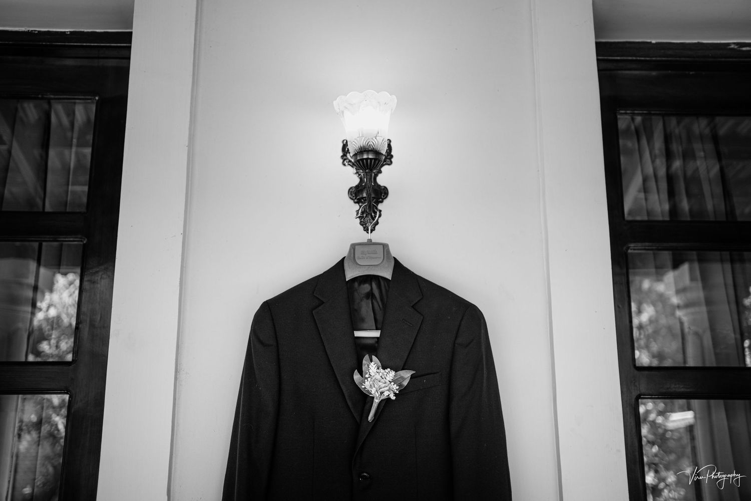 A black suit jacket hangs on a wooden hanger beneath a wall light fixture in black and white photography.