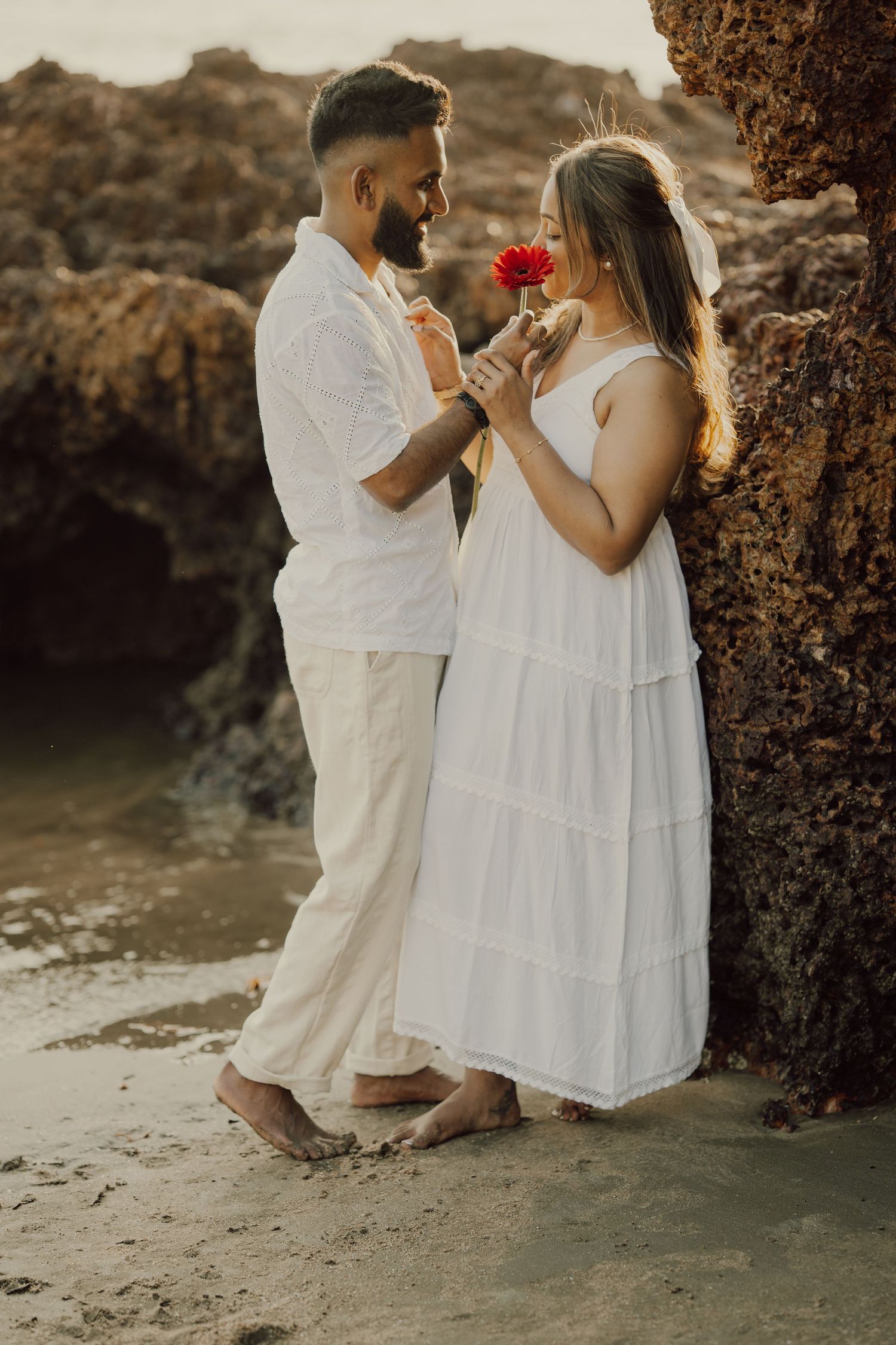 Romantic couple in white outfits sits on beach near rocky cliffs sharing tender moments with a red flower at sunset.