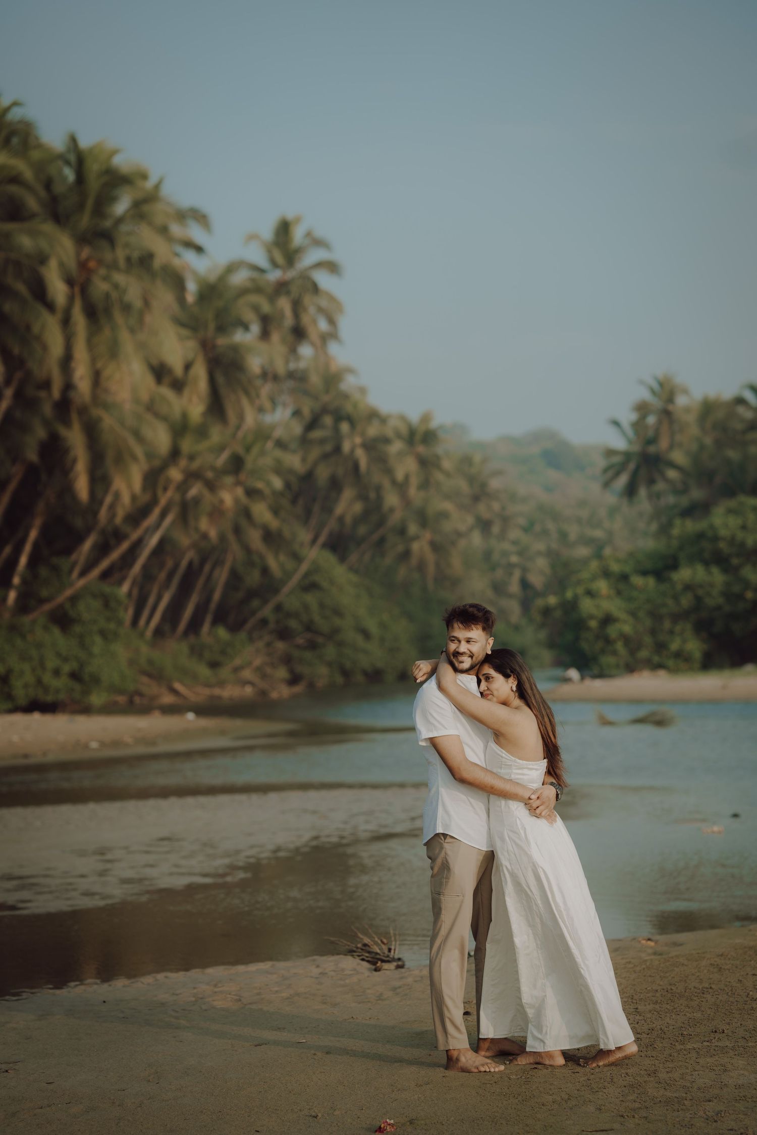 A couple shares an intimate moment on a tropical beach with palm trees and mountains in the background.