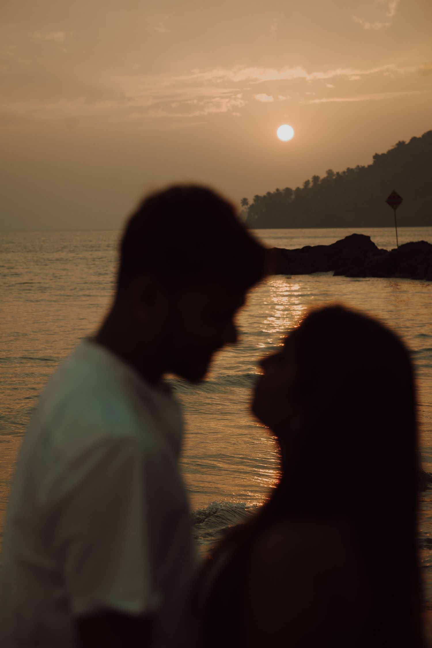 Silhouettes of two people against a sunset over water with mountains in the background creating a romantic scene.