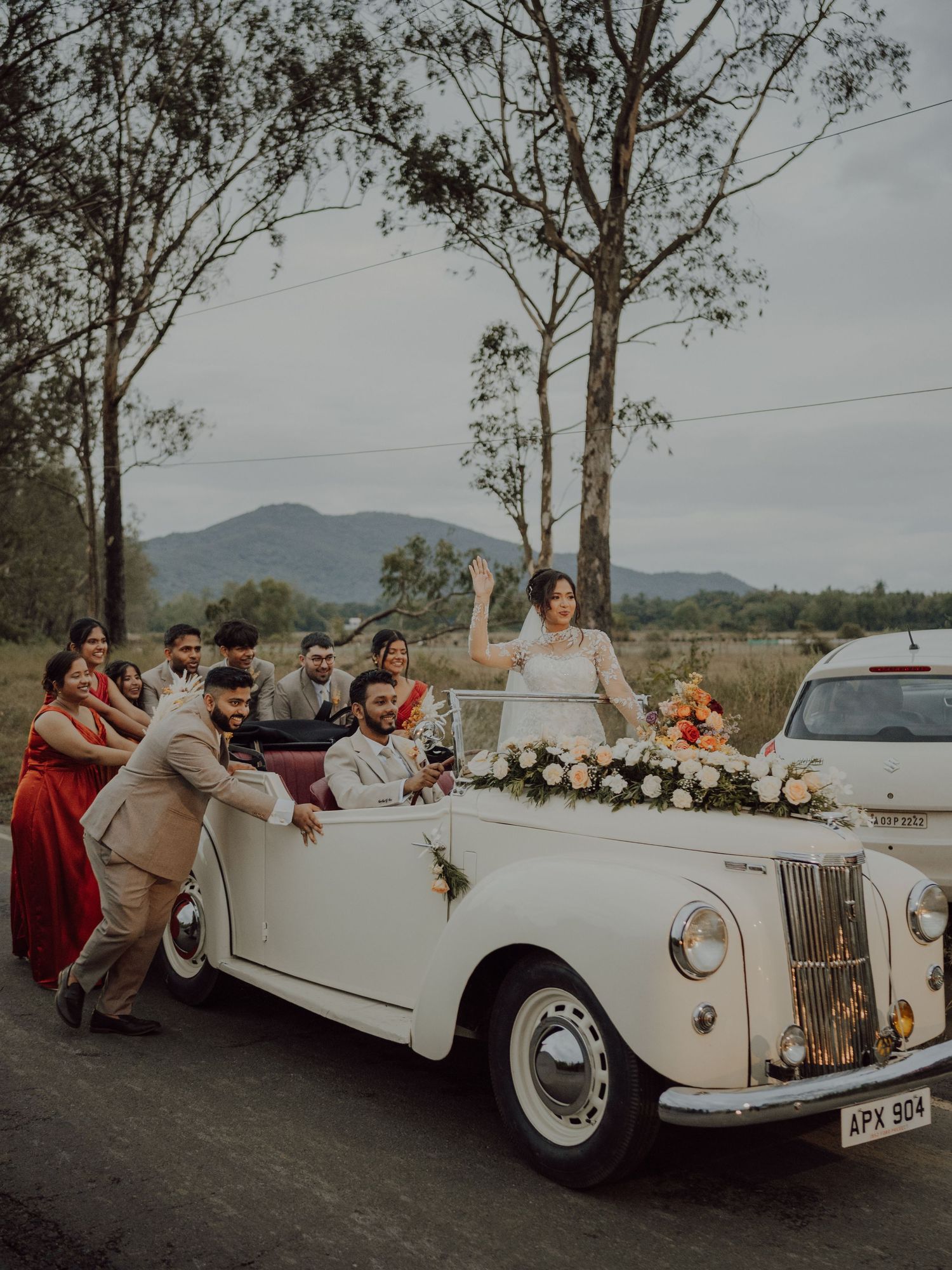 A bride stands in a white vintage convertible decorated with flowers as wedding guests gather around it on a country road.
