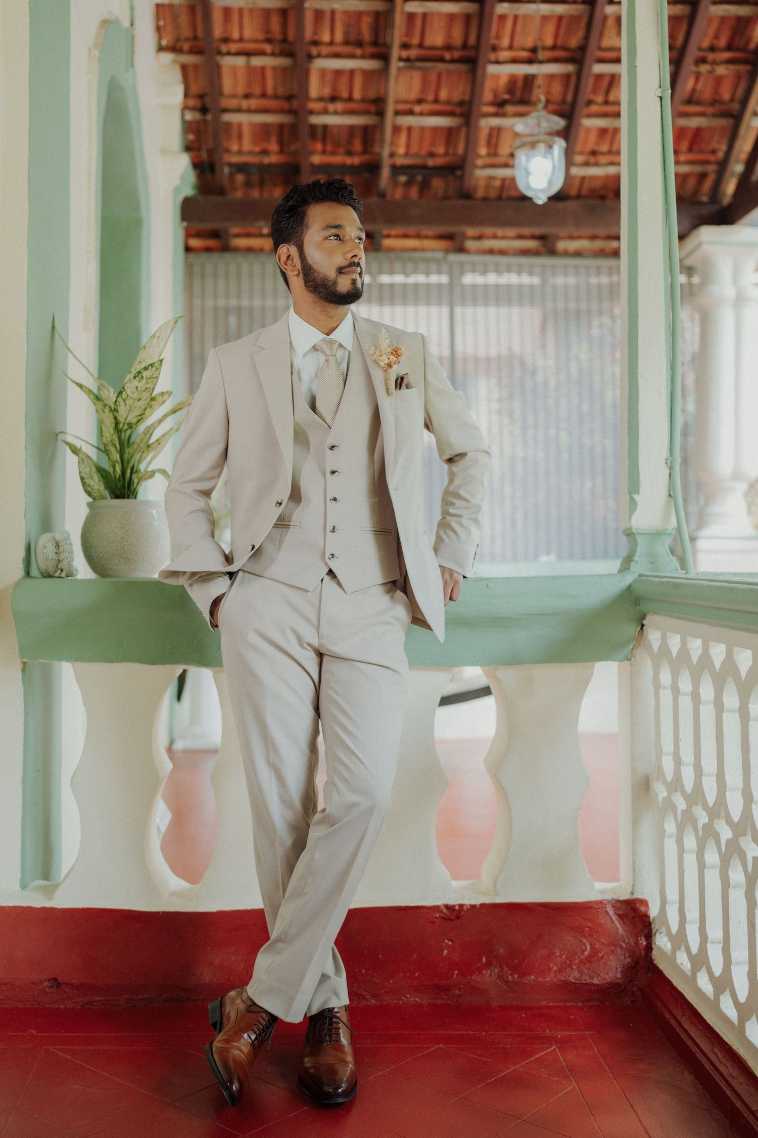 Groom in a beige three-piece suit and brown shoes leans on a white balustrade on a red-floored heritage veranda.