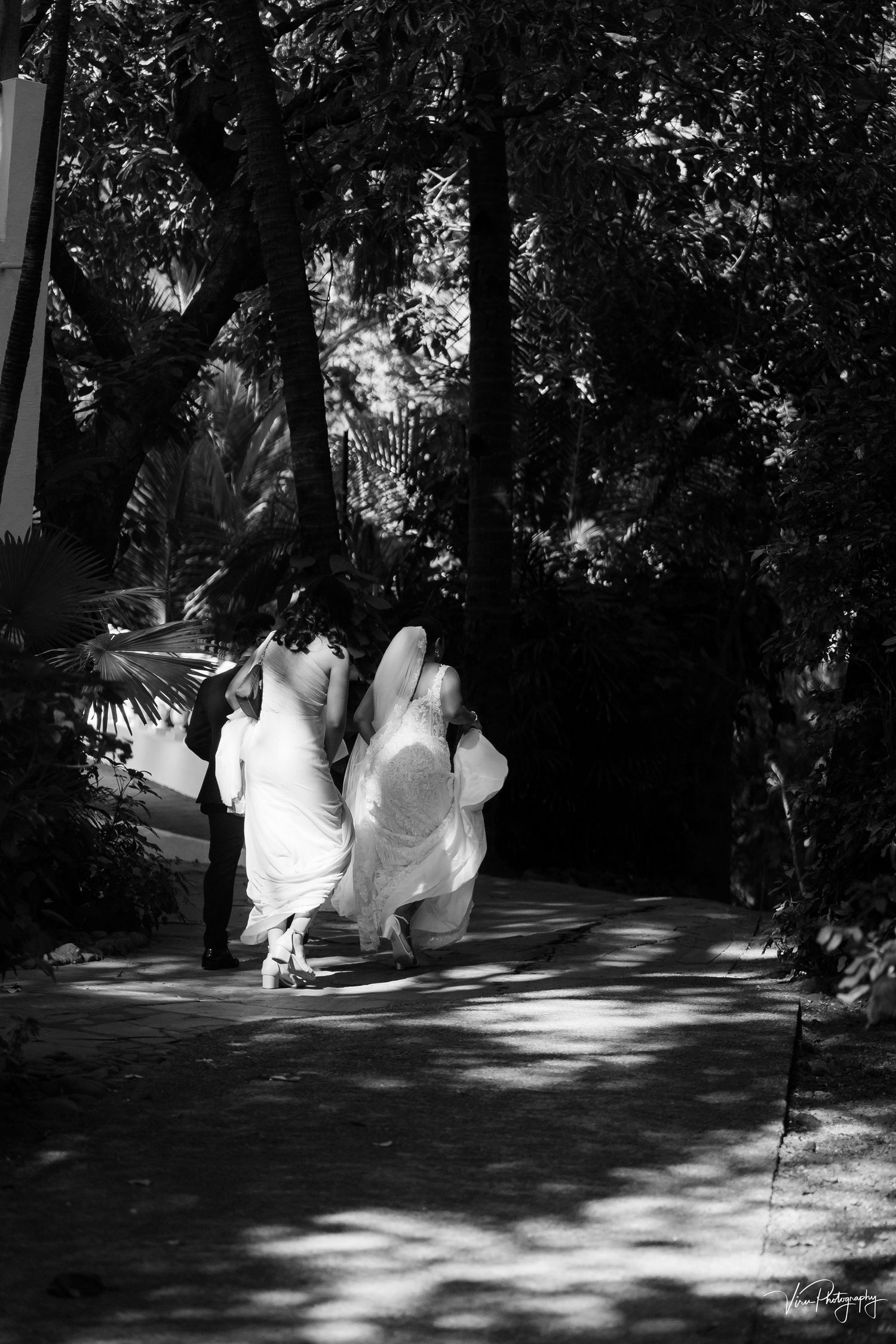 Two people in flowing white dresses walk together along a shaded garden path in black and white photography.