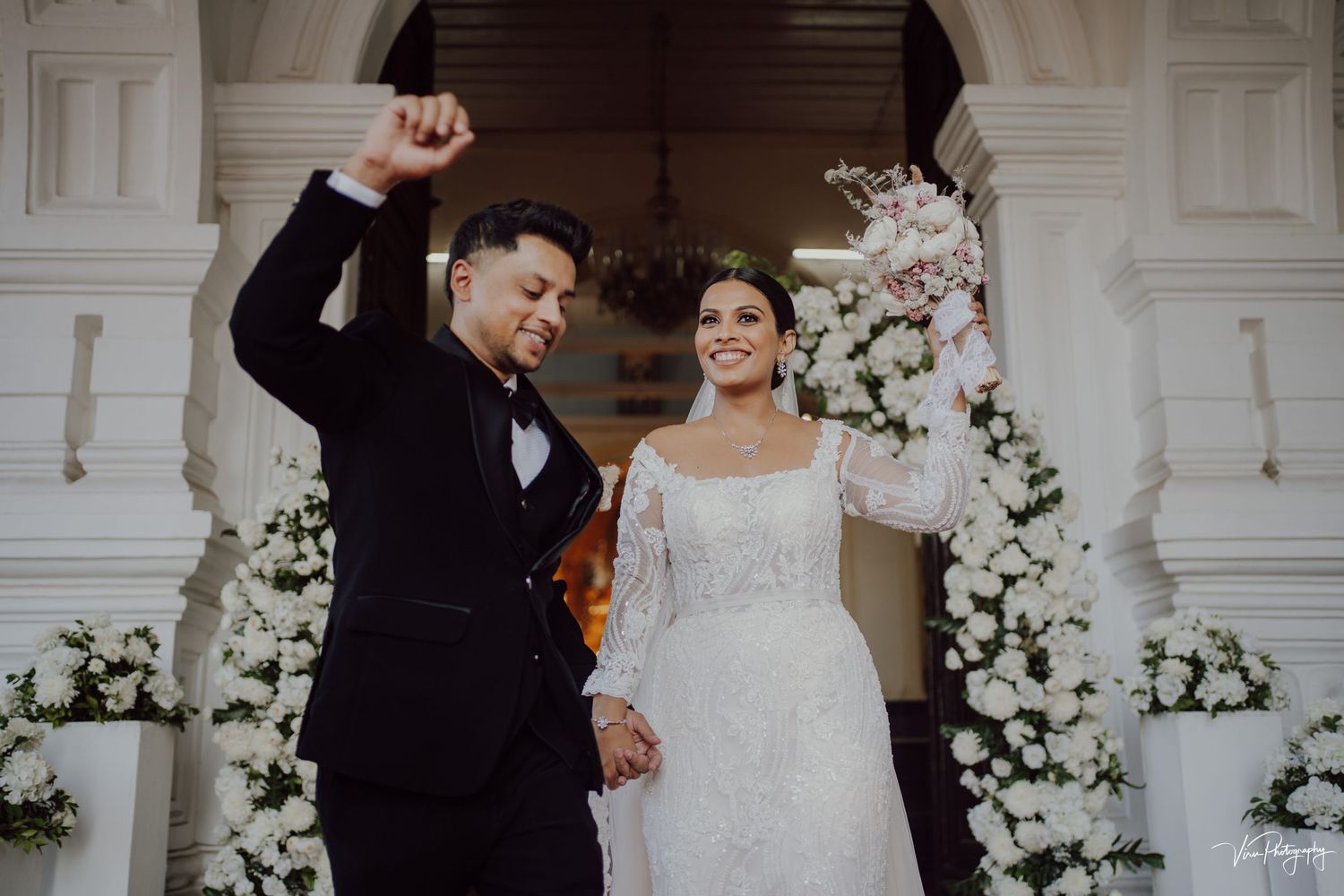 Newlyweds celebrate joyfully at their elegant wedding ceremony surrounded by white floral arrangements and archways.
