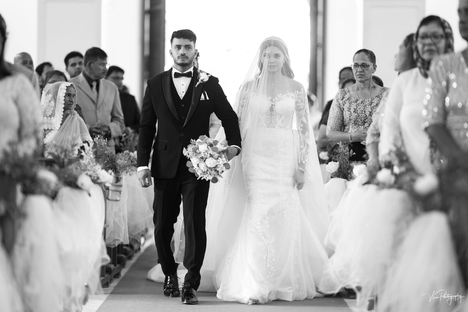 A bride and groom walk down the aisle together surrounded by guests in a black and white wedding photograph.