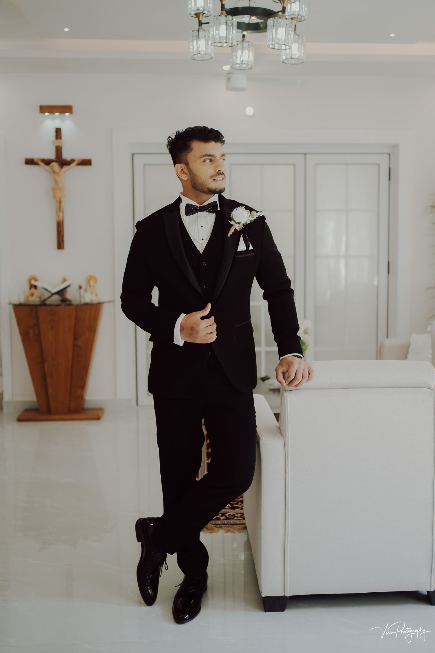 Elegant wedding attire featuring a black tuxedo with bow tie and boutonniere in a white chapel room with crucifix.