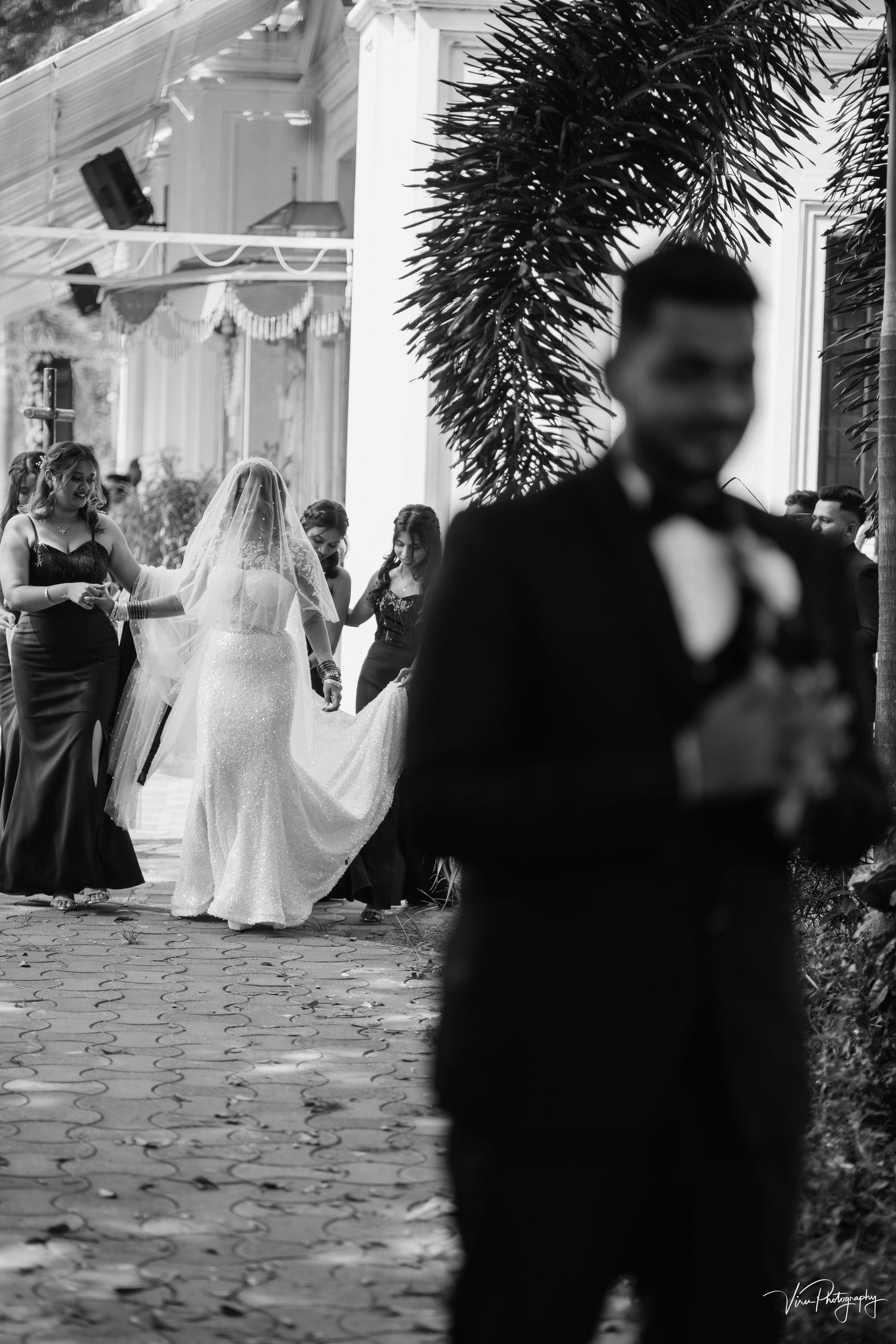A black and white wedding photo showing a bride in white dress and blurred figure in tuxedo during an outdoor ceremony.