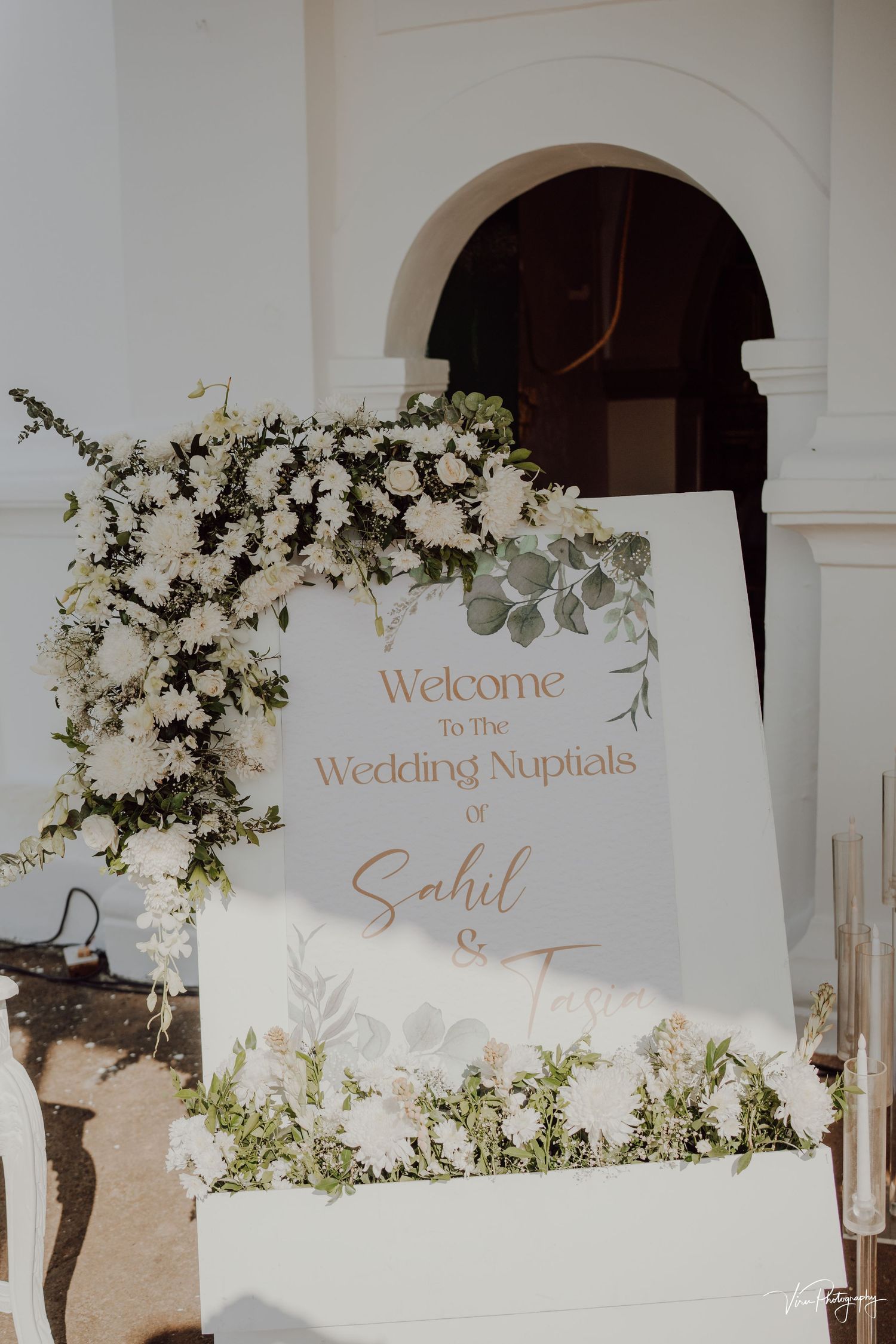 An elegant wedding welcome sign with white floral arrangements and eucalyptus branches decorating an archway entrance.