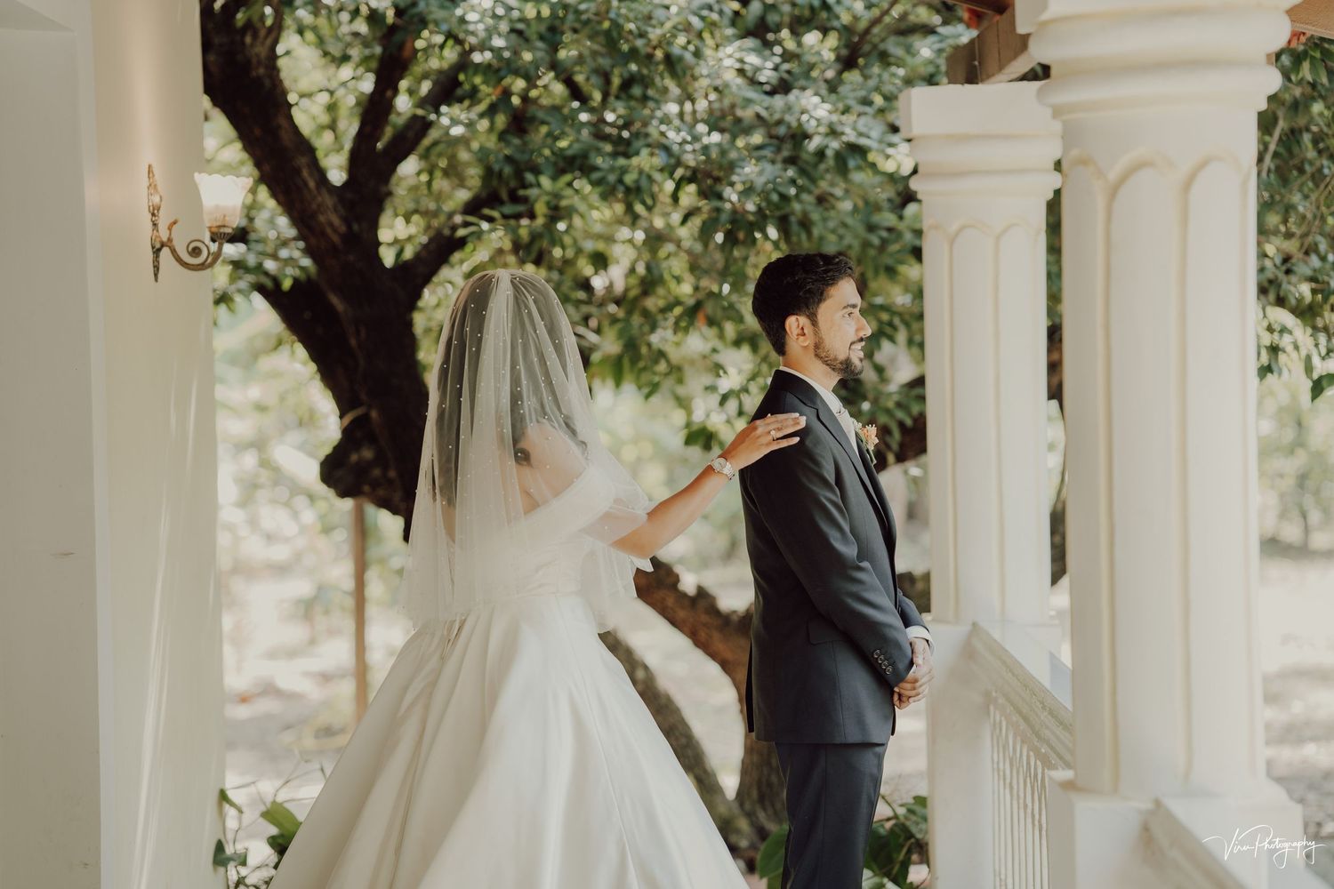 A bride and groom share an intimate moment on a white pillared porch surrounded by lush greenery and sunlight.