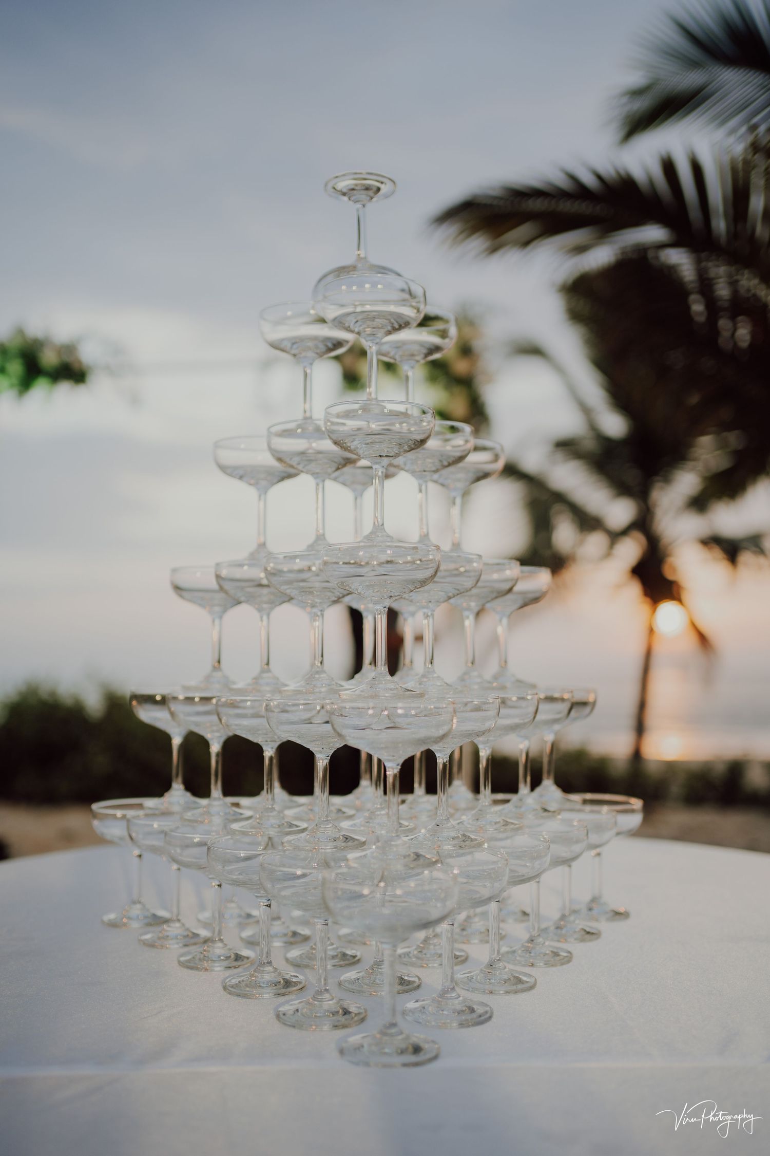 A champagne tower made of empty glasses stacked in tiers against a sunset sky with palm trees silhouetted in the background.