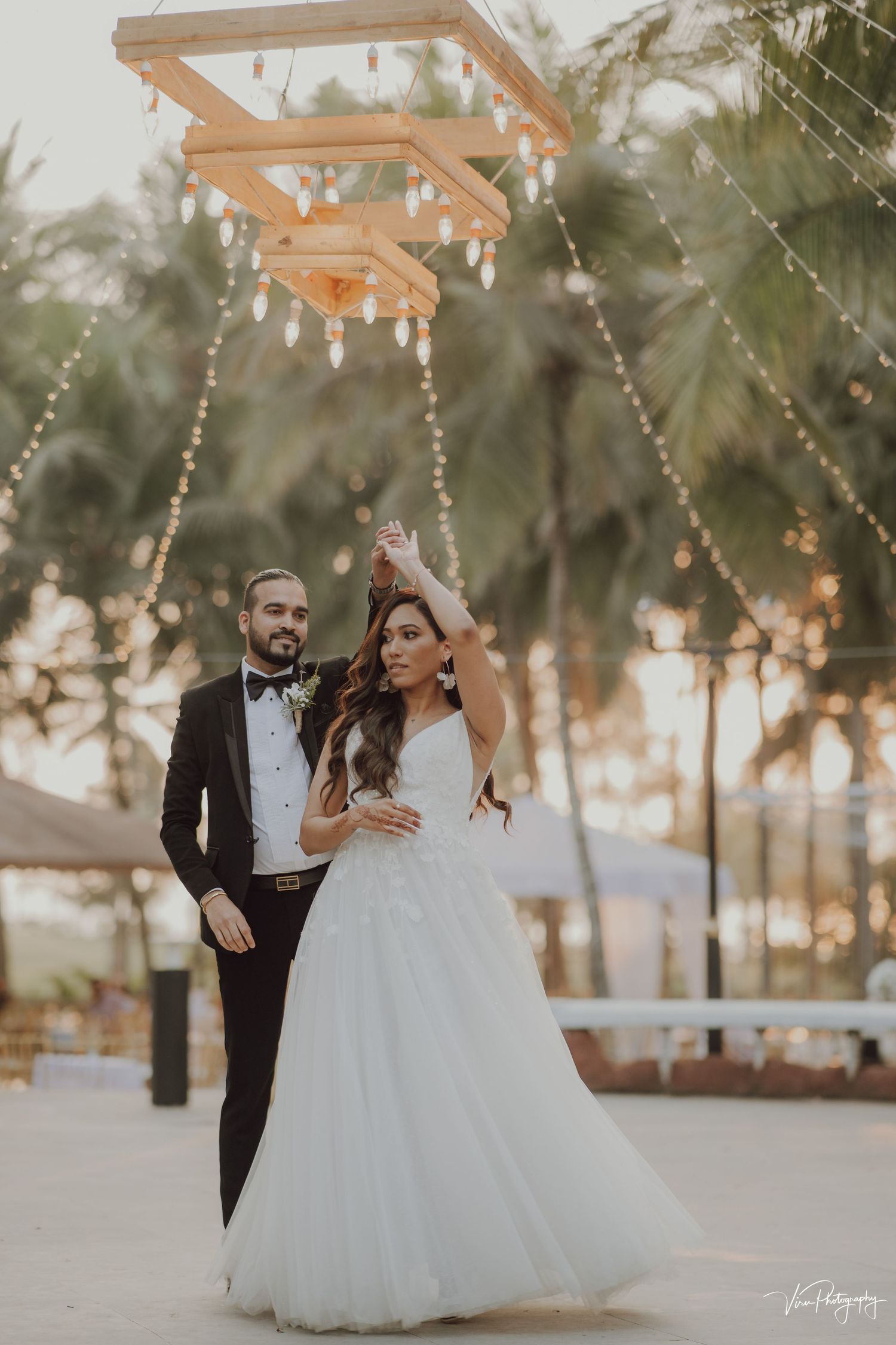 Couple in formal wedding attire dancing underneath a wooden pergola with hanging lights at sunset and palm trees in background.