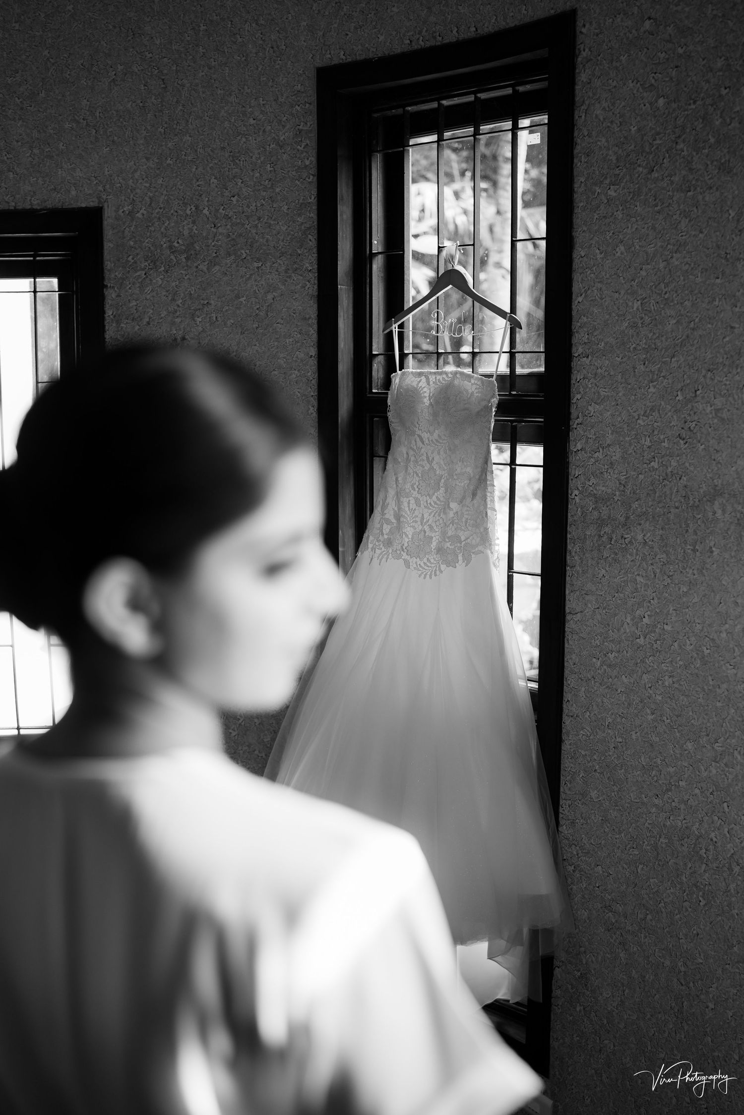 A white wedding dress hangs in silhouette against a window frame as a figure in the foreground stands in soft focus.