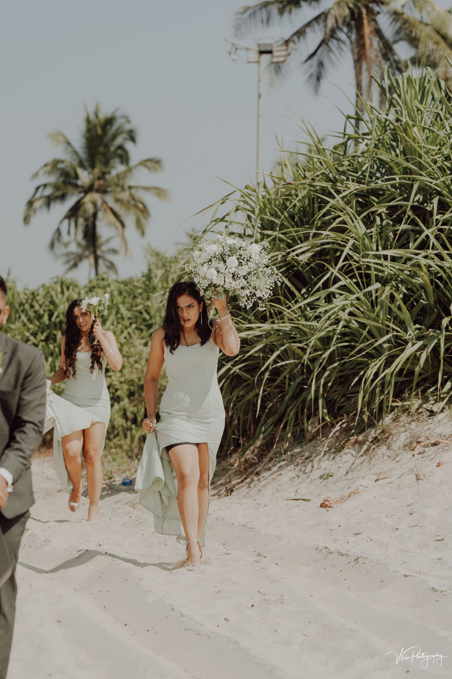 People walking on a sandy beach path surrounded by tropical palm trees and lush vegetation on a sunny day.