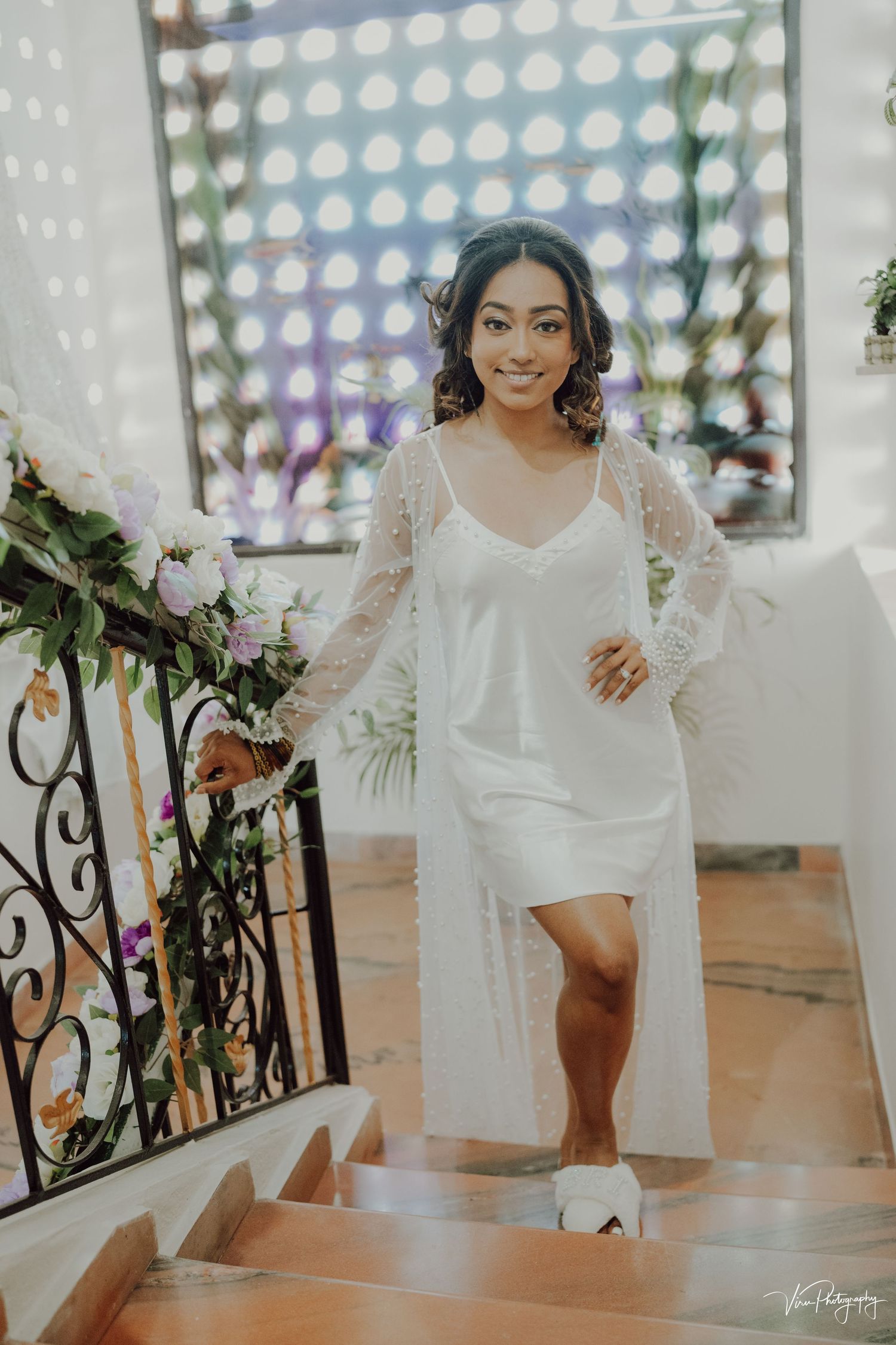 A woman in white silk robe and slip dress descends stairs in a sunlit room with dotted wall pattern.