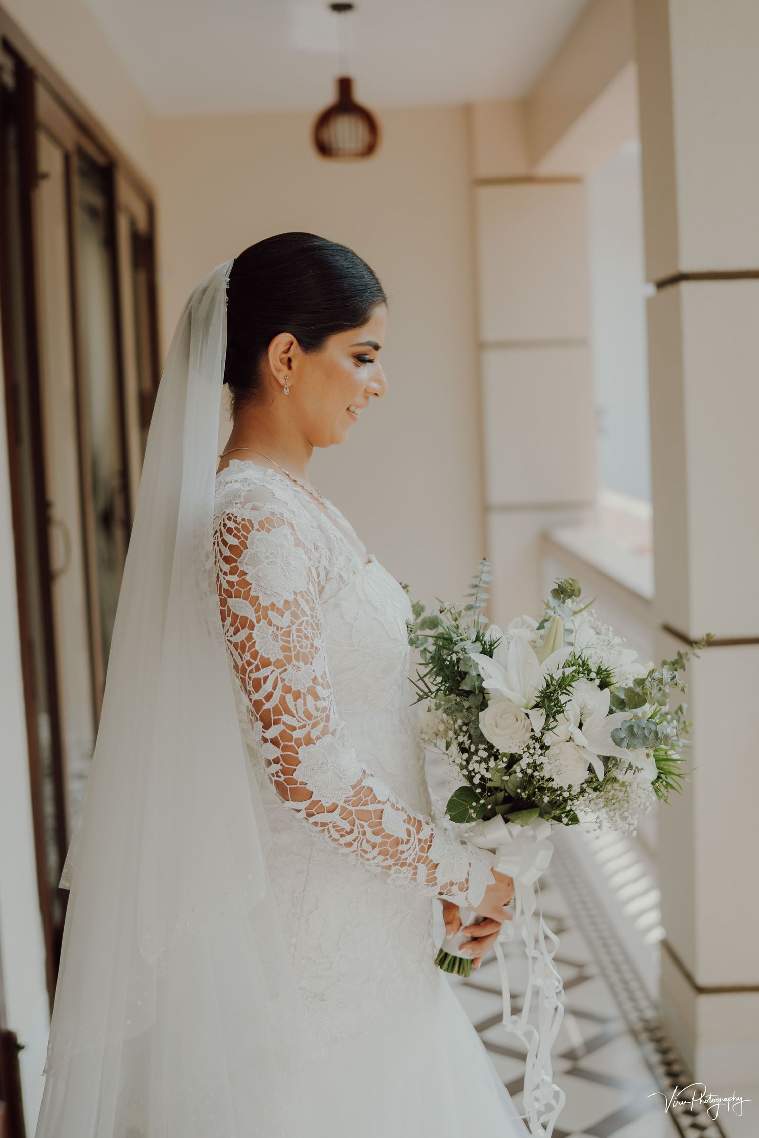 A bride in a long-sleeved white lace wedding gown holds an elegant white and green bouquet on a classic veranda.