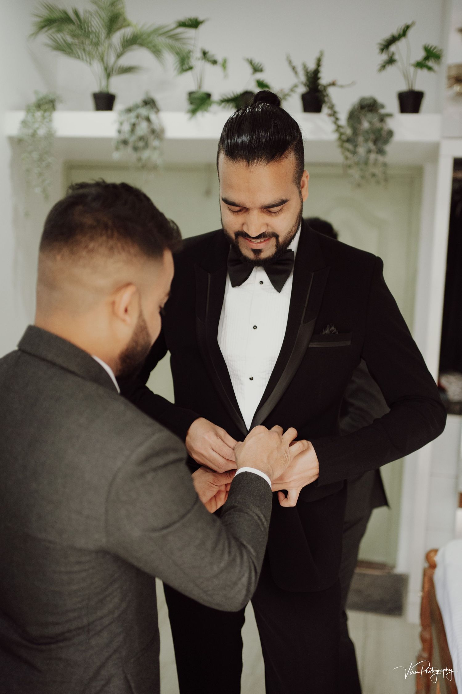 Groomsman helping adjust the bowtie and tuxedo of smiling person at indoor wedding venue with potted plants on shelf.