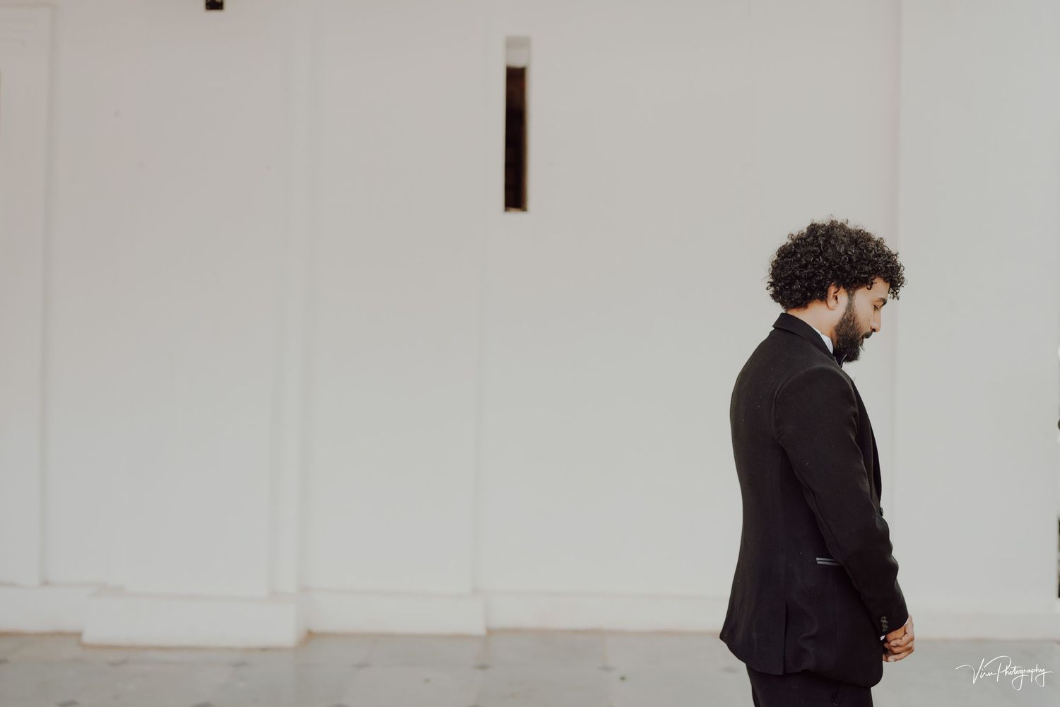 A person in a black suit stands against a white wall in a minimalist setting.