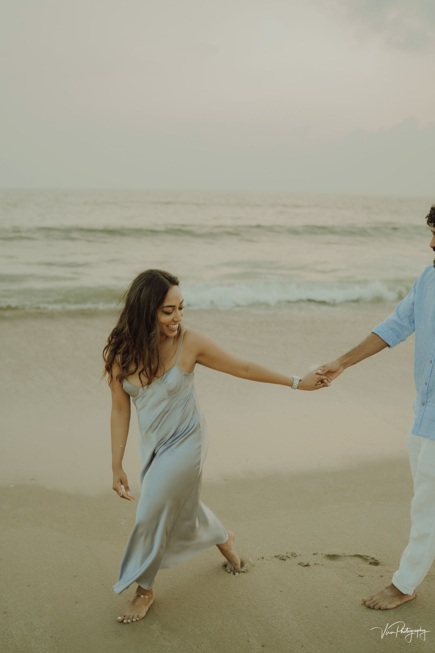 A couple holds hands and walks along a misty beach shore, with the woman wearing a flowing light blue dress.