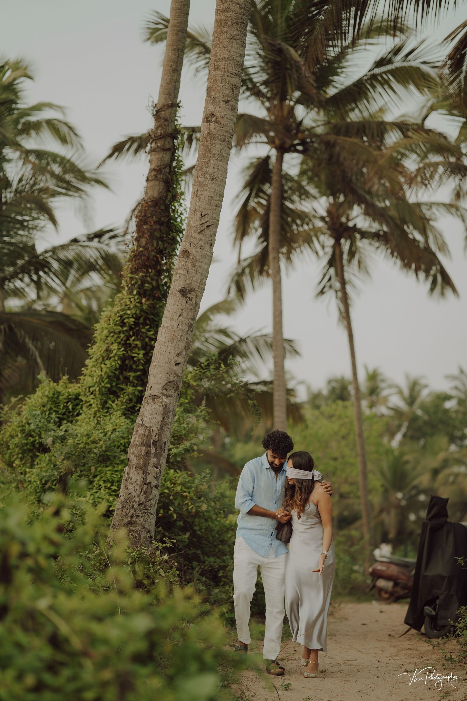 A couple walks hand in hand along a tropical beach path lined with tall swaying palm trees and lush greenery.