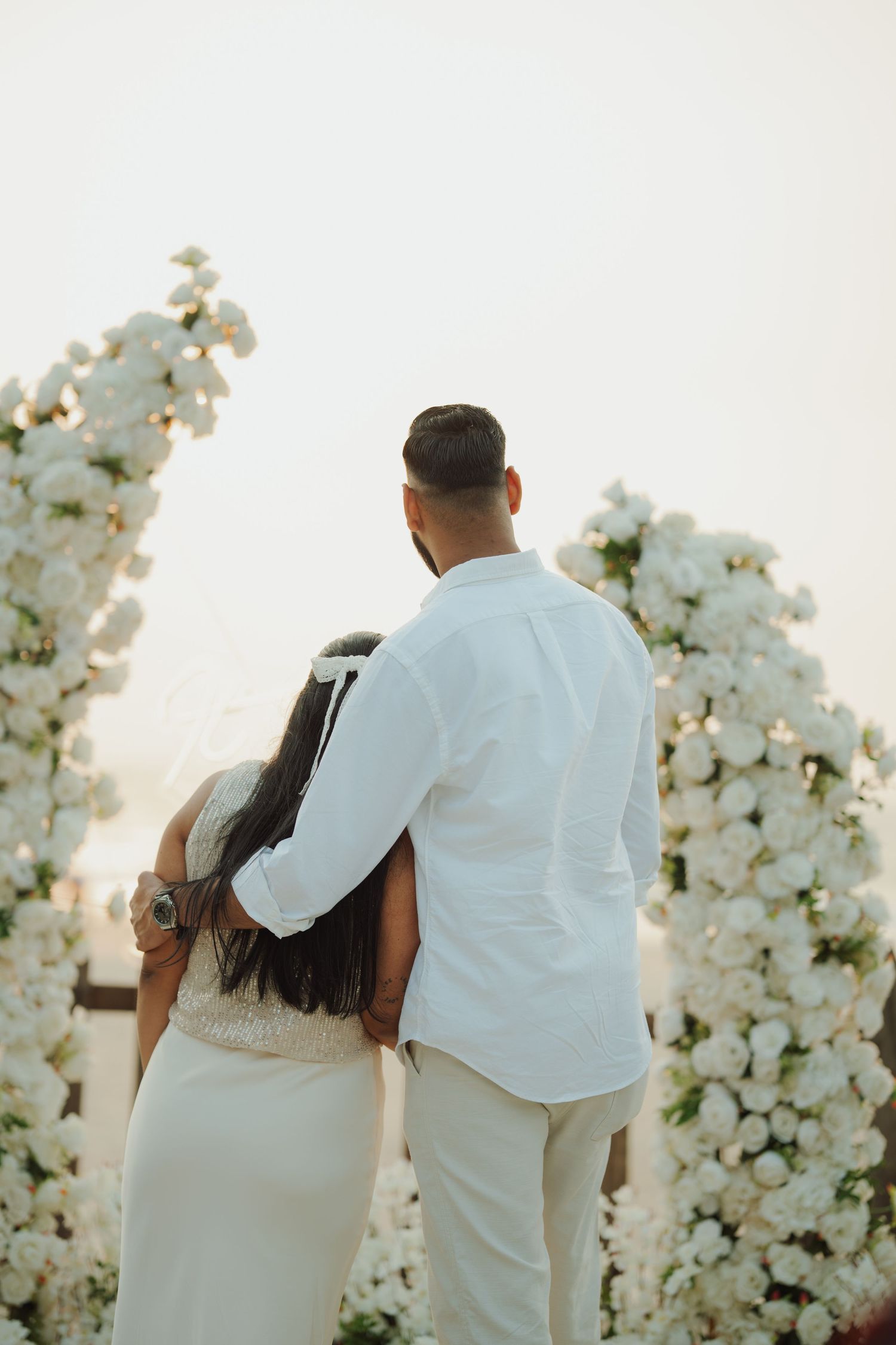 A couple in white attire embraces before a floral arch adorned with white roses at an outdoor wedding ceremony.