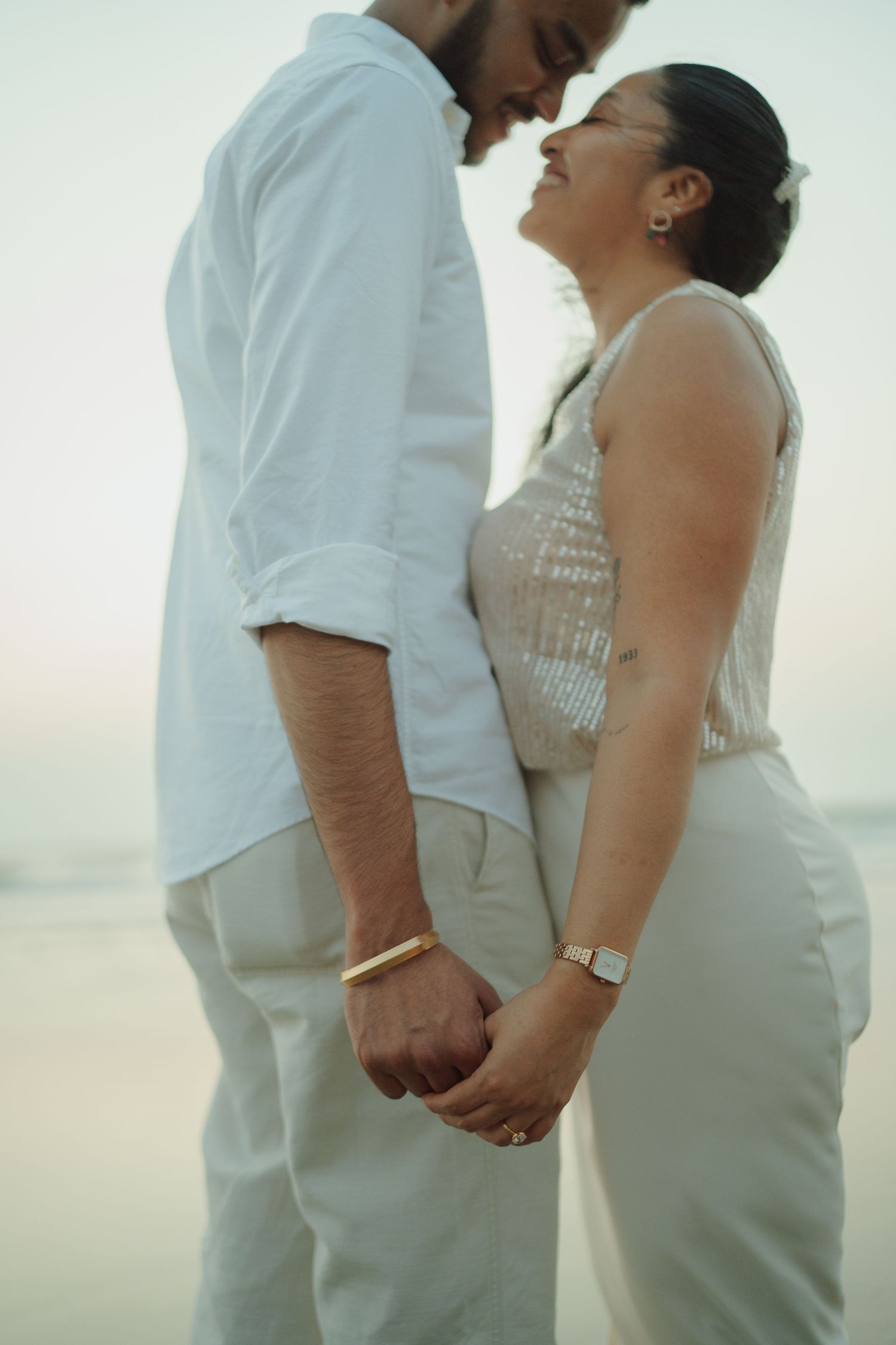 Couple holding hands on beach at sunset, wearing white outfits, sharing a romantic moment together.