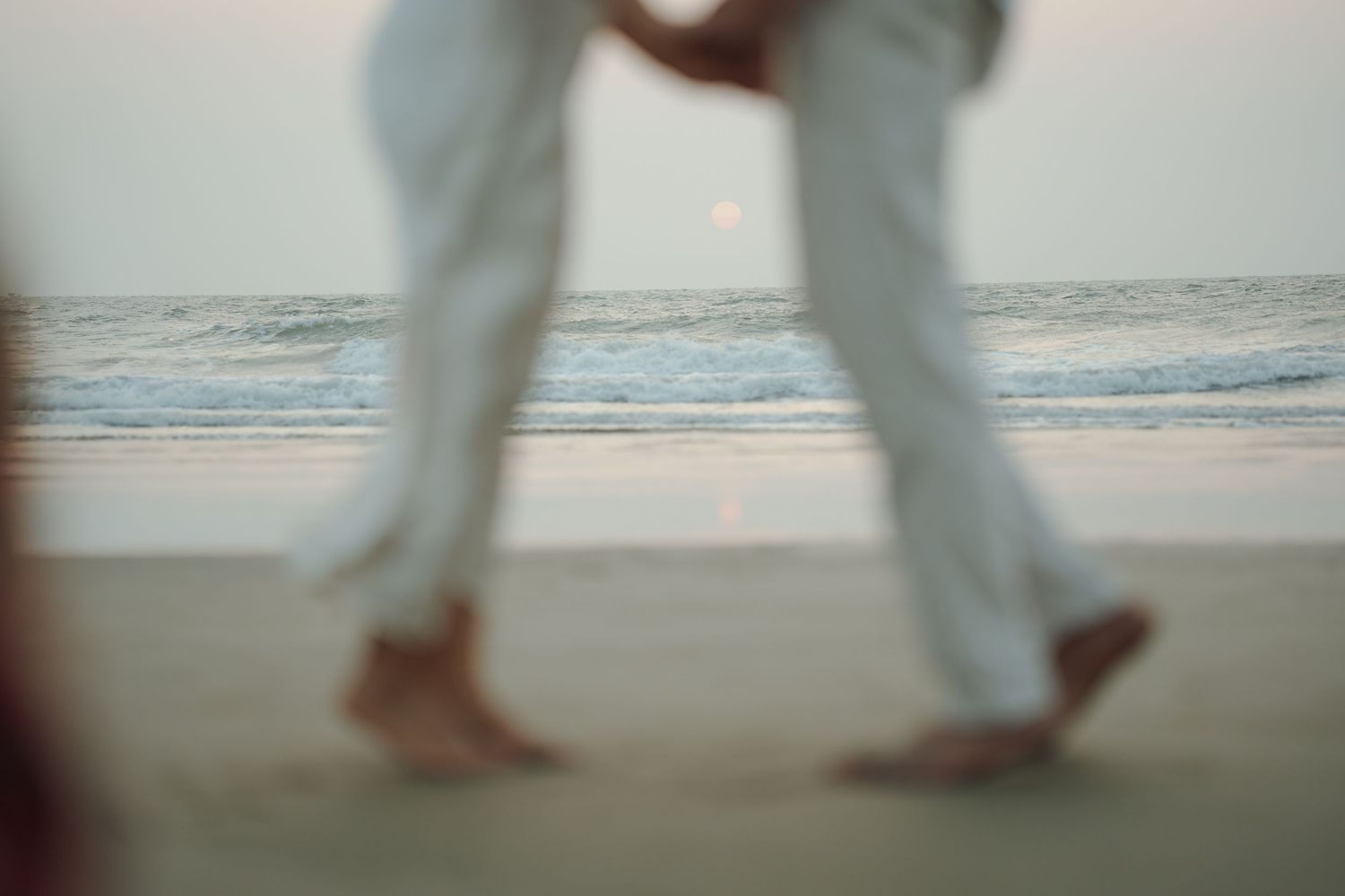 Barefoot couple in white pants dancing on a sandy beach at sunset with ocean waves in the background.