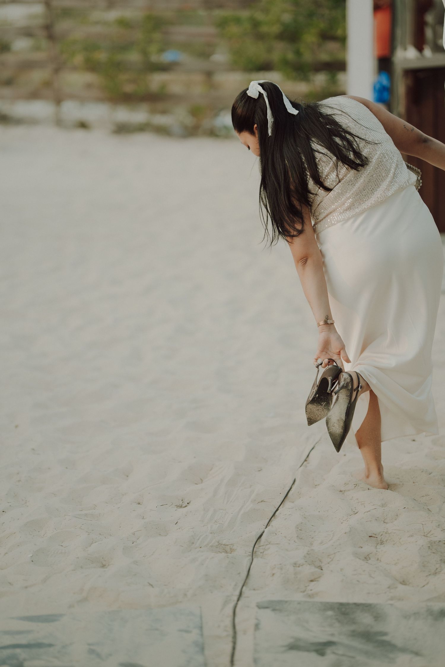 Woman in white dress holding heels on sandy beach, dark hair with white bow accessory, bending down barefoot.