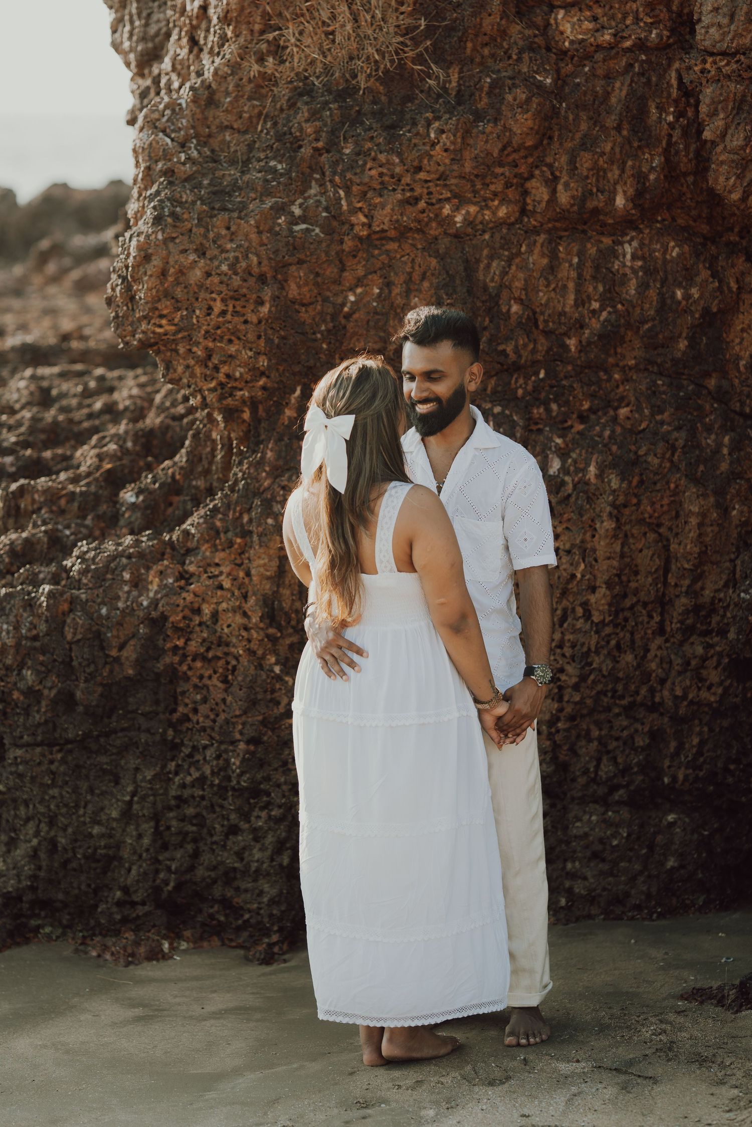 A couple in white outfits poses together against a rustic stone wall during a romantic outdoor photoshoot.
