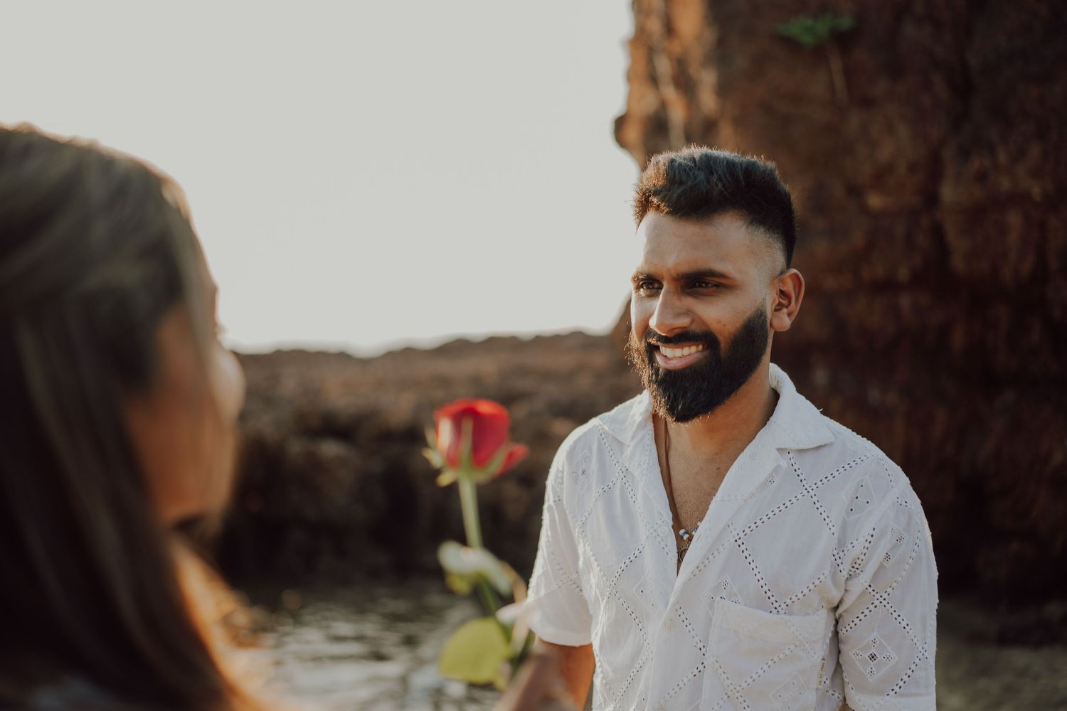 A couple in white outfits shares an intimate moment at sunset on a rocky beach landscape.