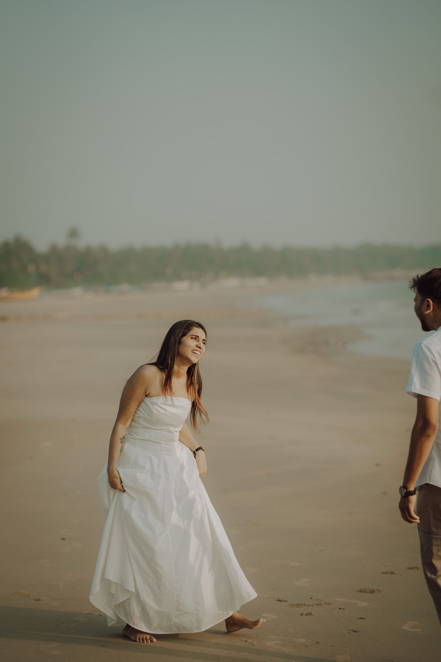 A woman in a flowing white dress walks gracefully along a sandy beach shoreline.