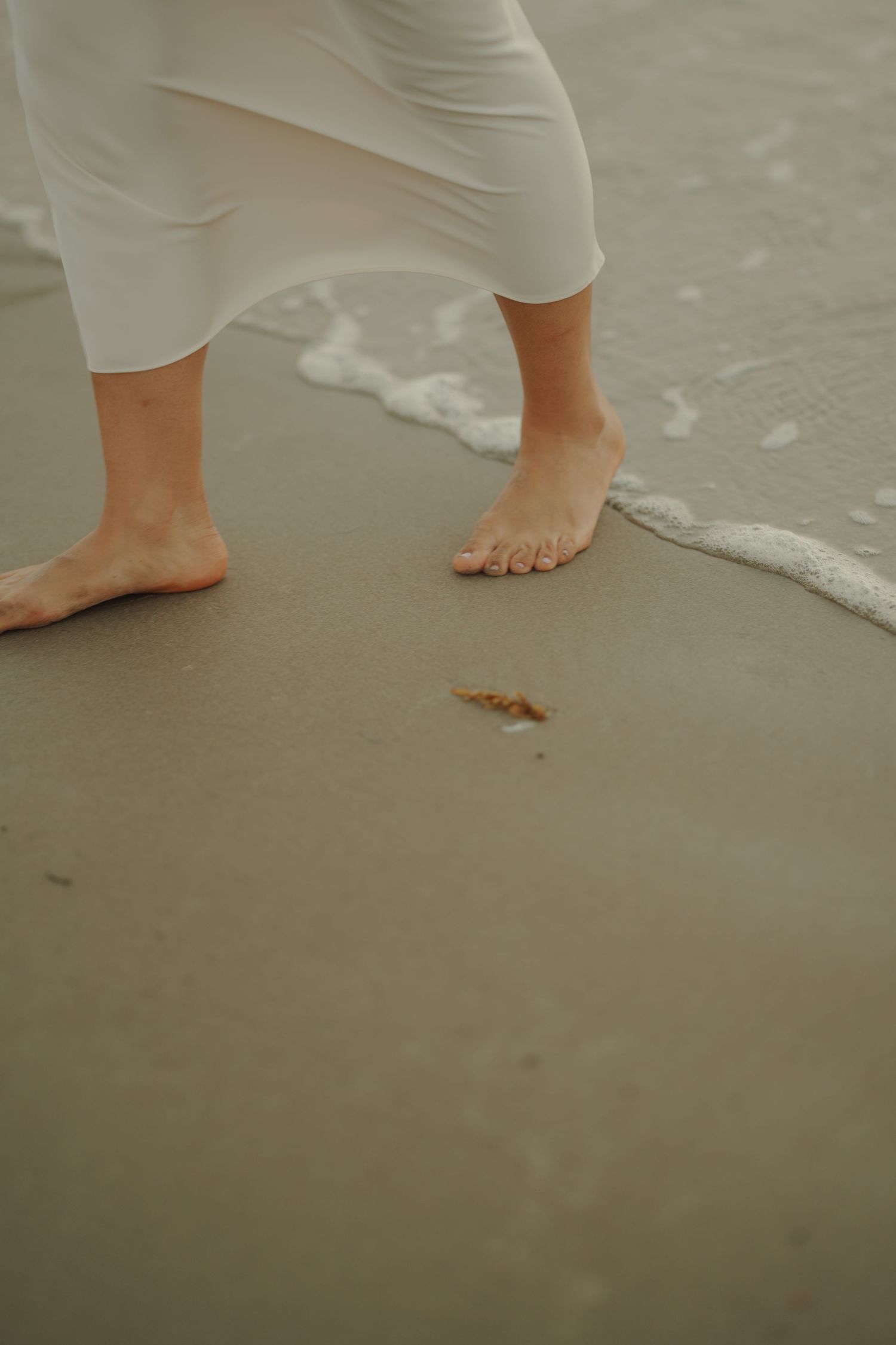 Barefoot person in white dress walking on wet sandy beach with ocean waves washing ashore.
