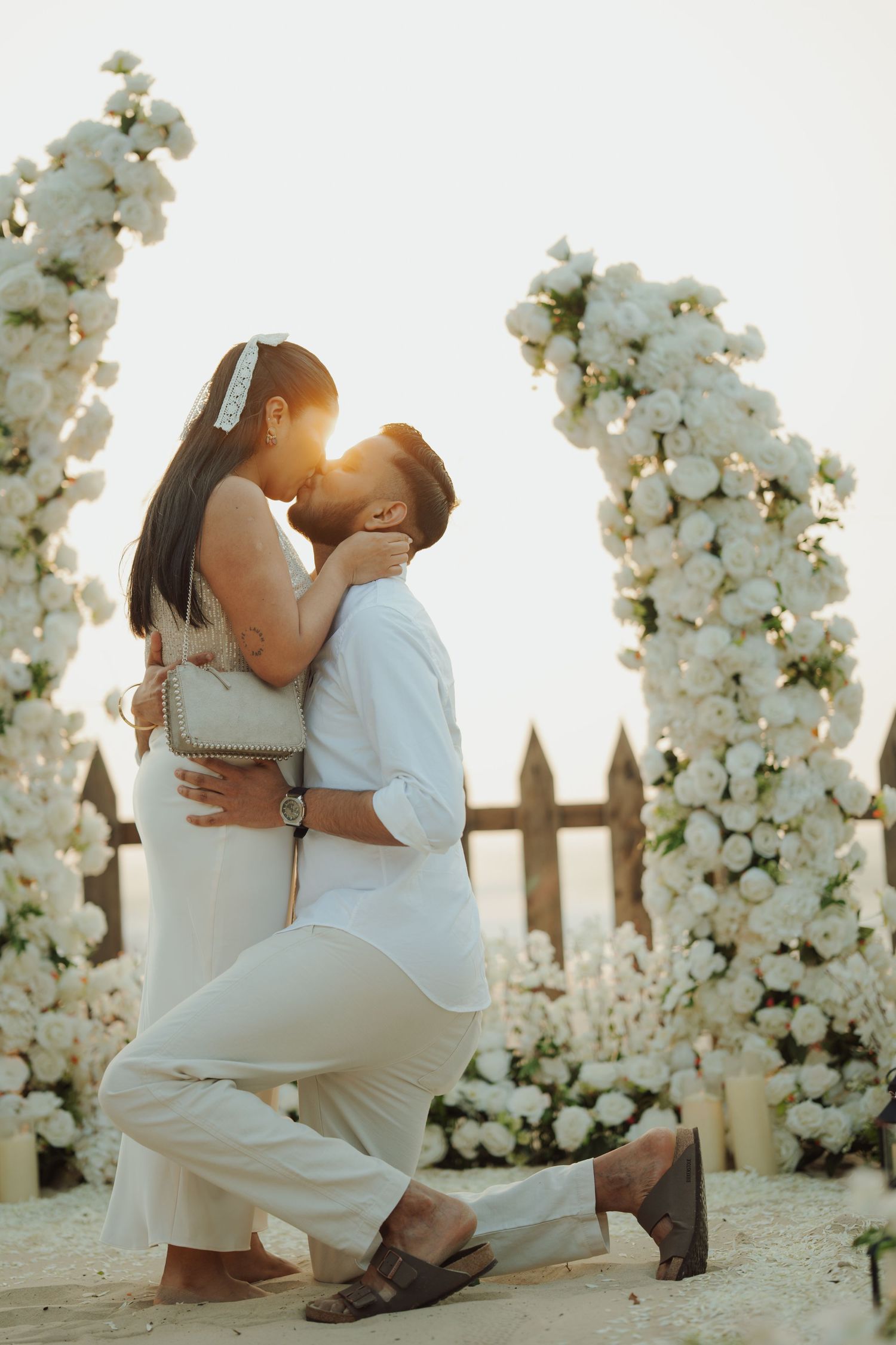 Couple kissing during golden hour proposal surrounded by white floral arches, man on one knee in white outfit outdoors.