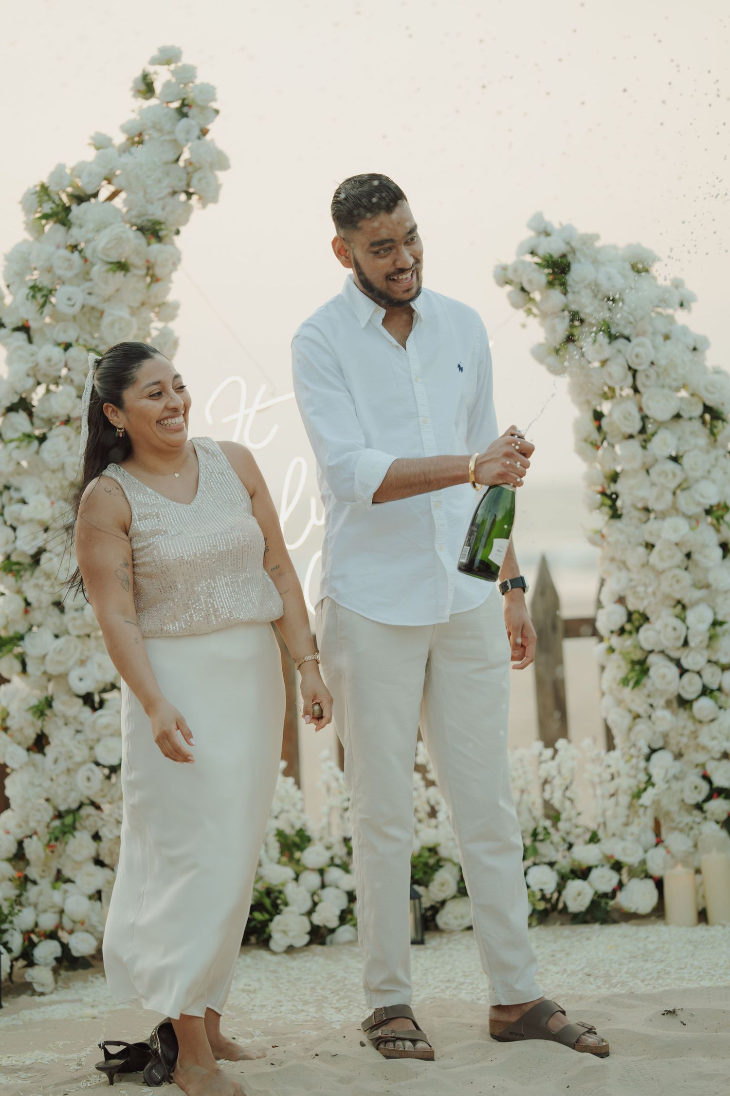 Couple celebrating engagement on beach with champagne, white floral arch backdrop, wearing matching white outfits.