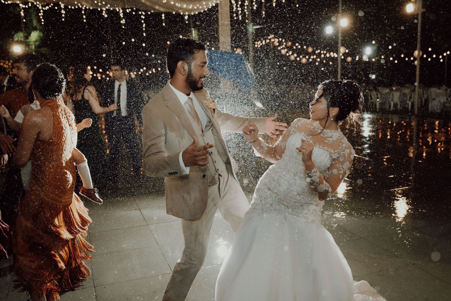 A bride and groom dance in a spray of water droplets under string lights while wedding guests celebrate around them at night.