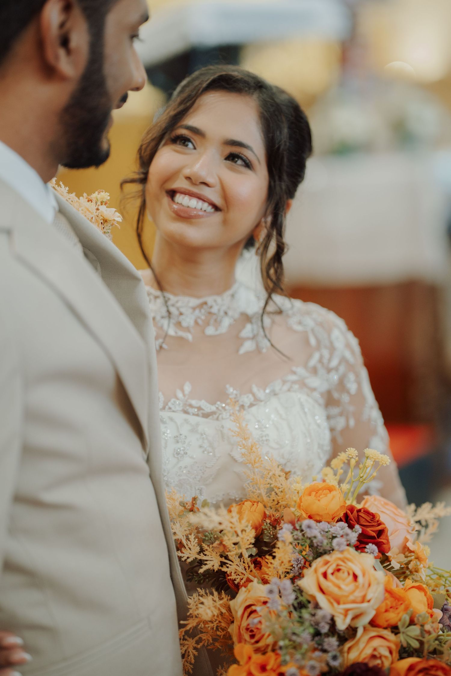 A smiling bride in a lace wedding gown holds an orange and peach rose bouquet while gazing at her groom in a beige suit.