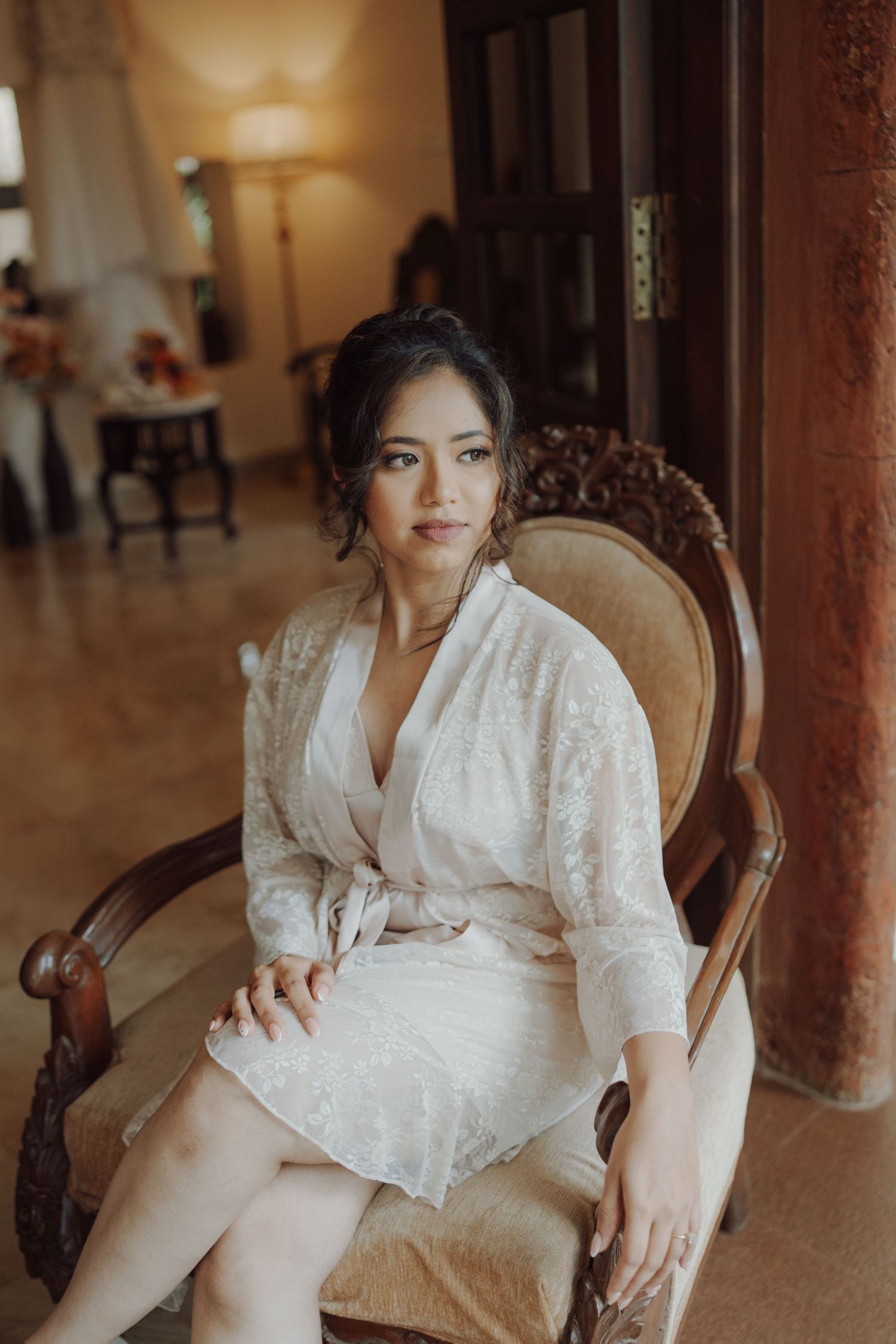 A bride in a white lace robe sits gracefully in an ornate carved wooden chair in an elegant interior before her wedding.