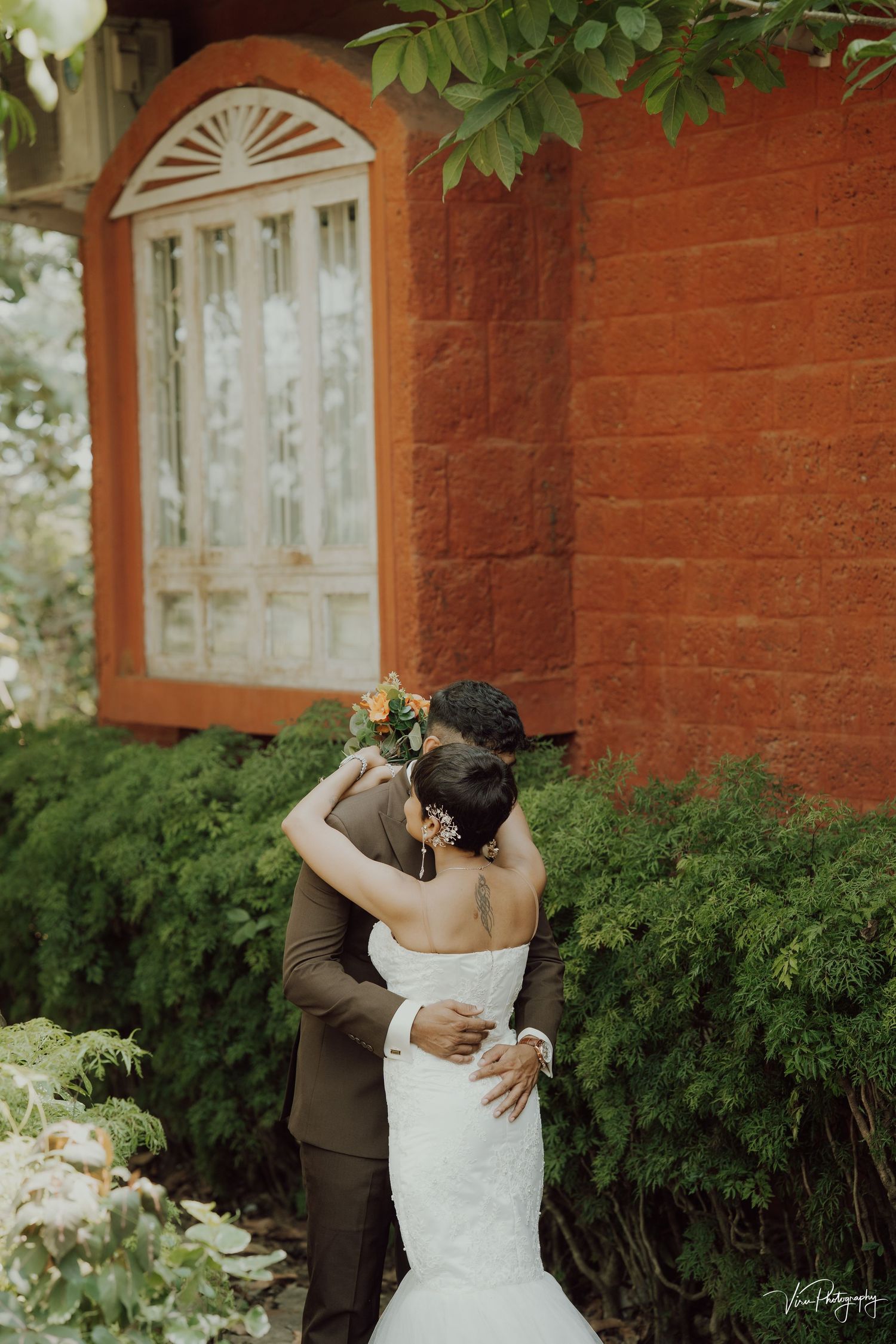 A romantic embrace at golden hour in front of a rustic red brick wall with greenery.