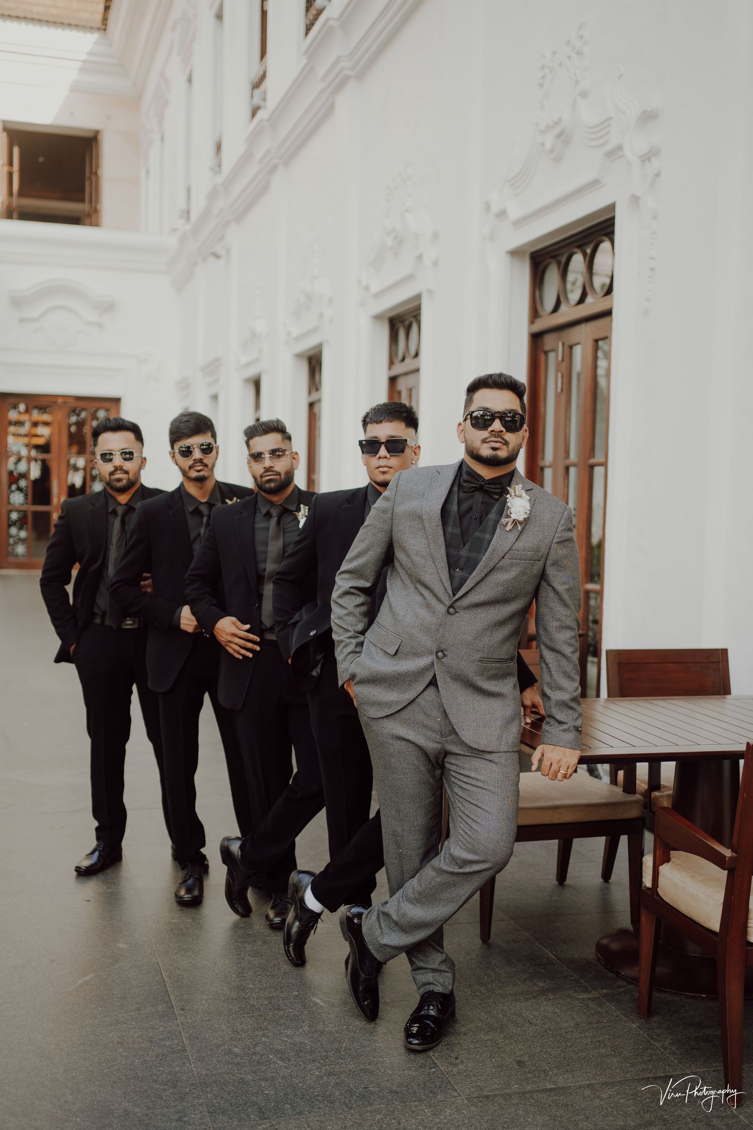 Stylishly dressed groomsmen in black and gray suits pose together in an elegant indoor venue with white walls.
