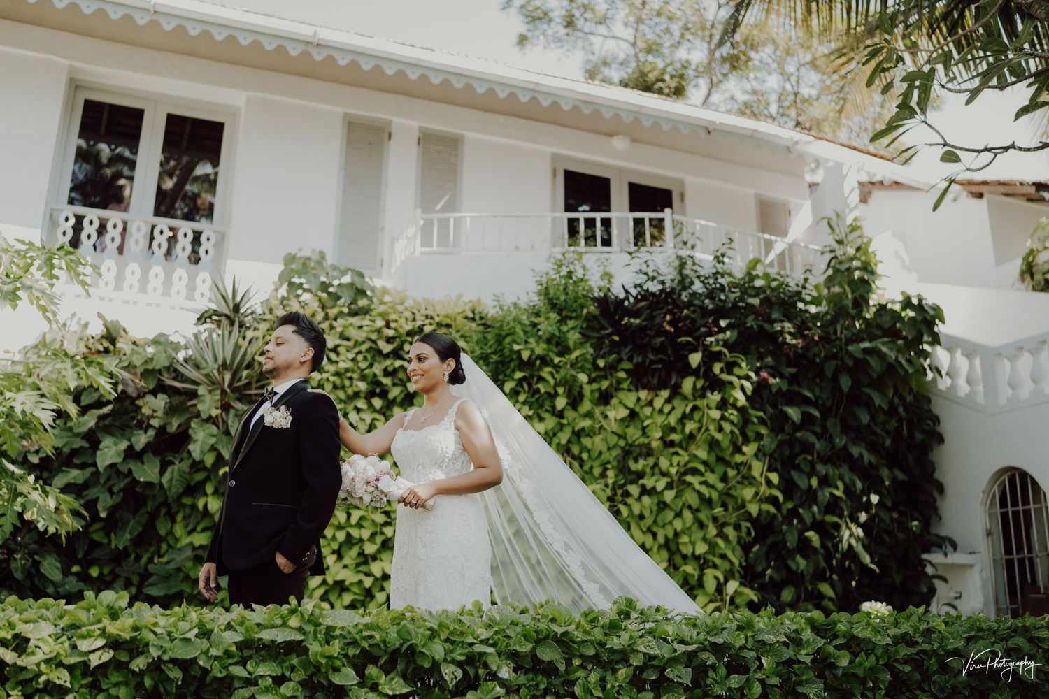 Couple in formal wedding attire poses in front of white colonial style building with lush garden landscaping.