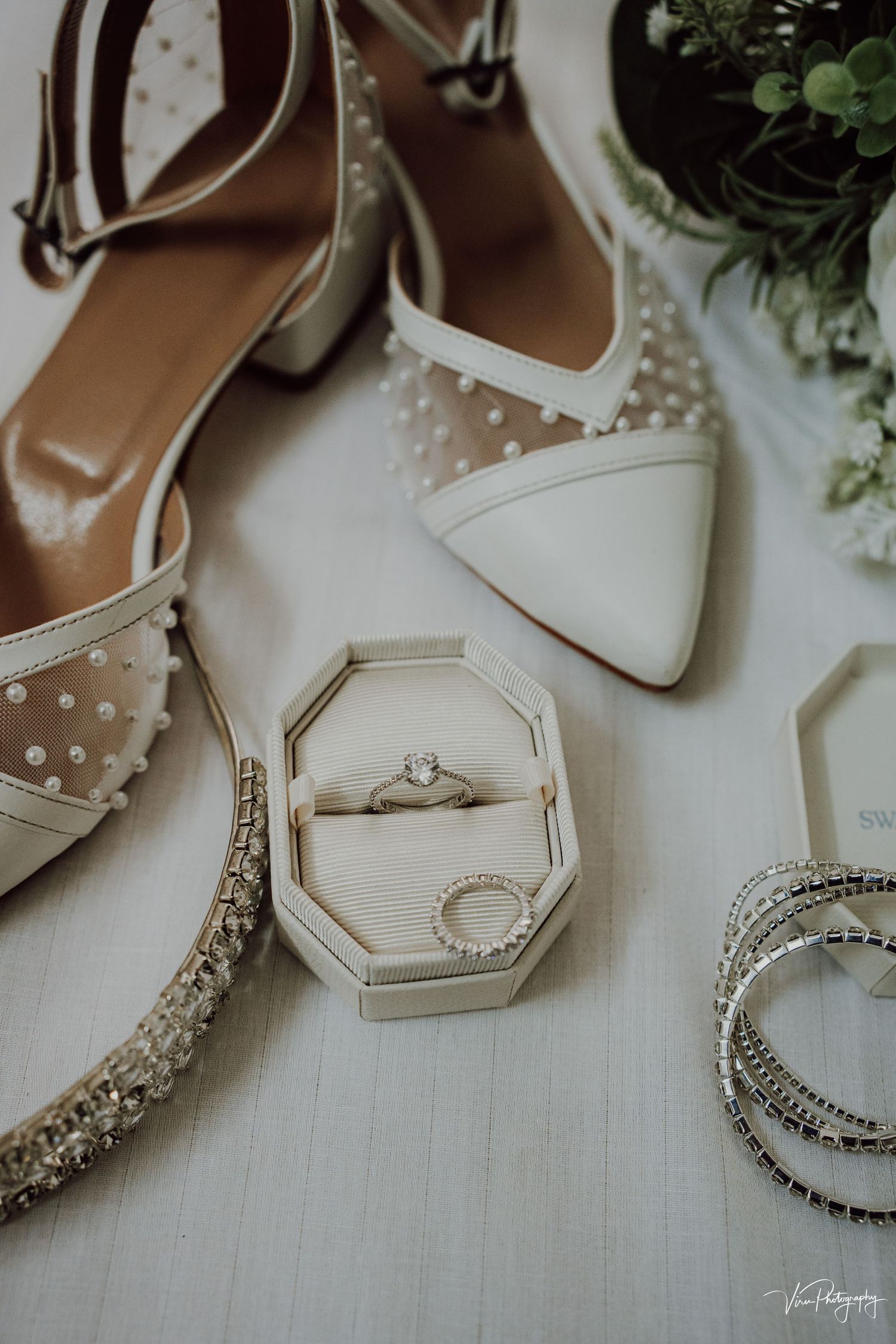 Elegant white bridal shoes with mesh polka dots displayed next to a ring box and sparkly bracelet accessories.